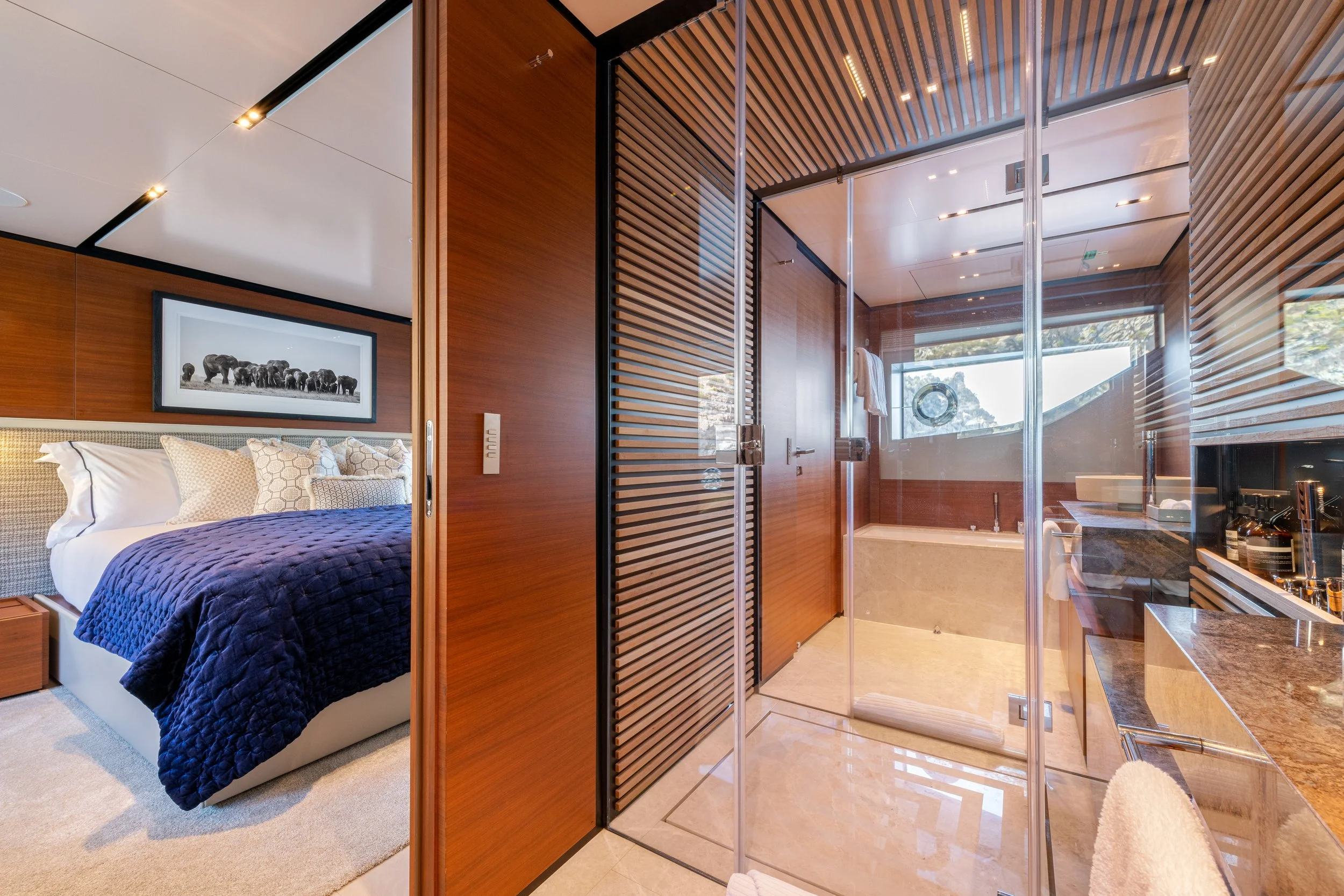 A bedroom with a bed featuring a navy quilt and patterned pillows, and a bathroom with a walk-in shower and a large bathtub, separated by a glass wall. The room has wood paneling and modern decor.