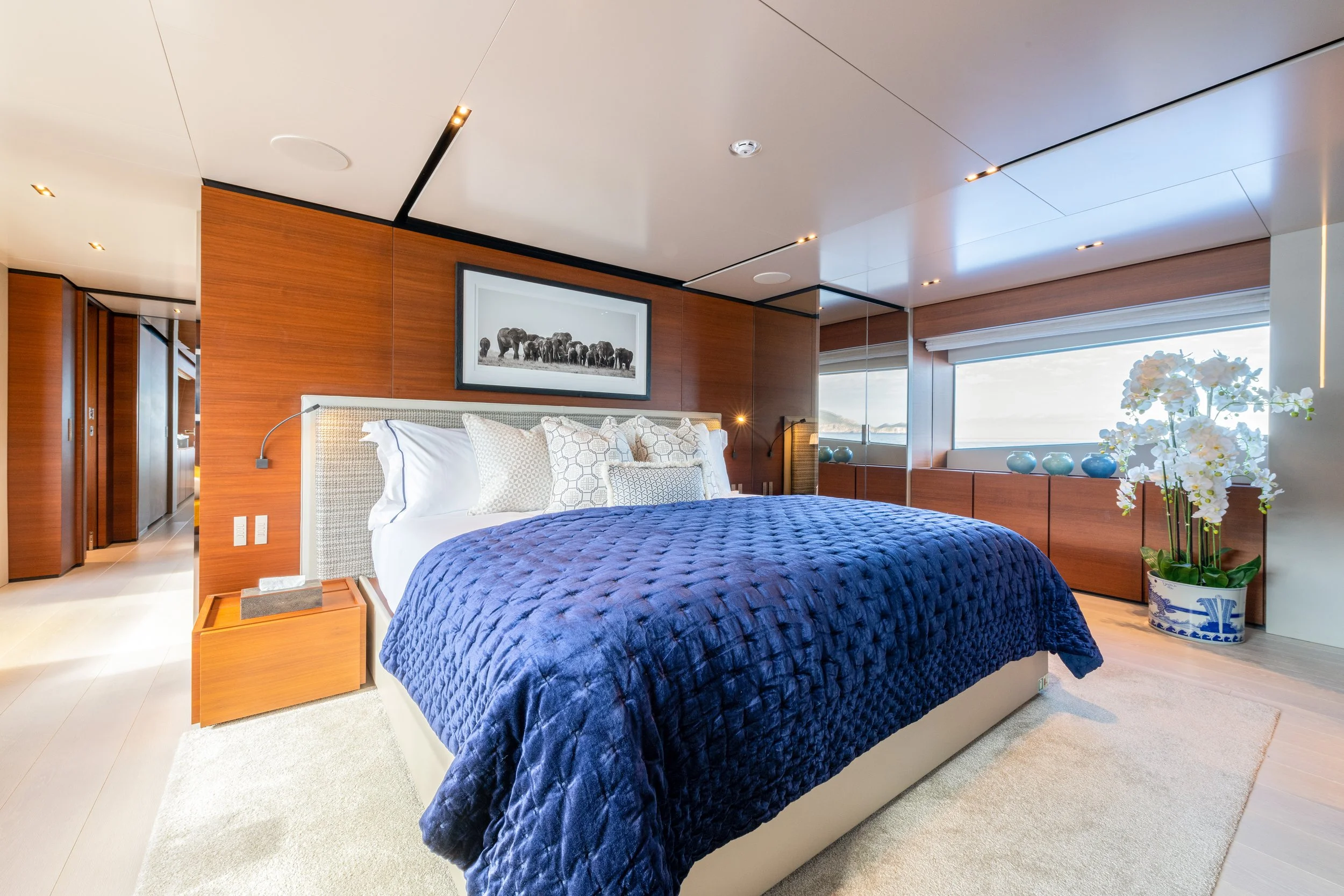 Modern bedroom with wooden wall panels, a large bed with white and decorative pillows, a blue quilt, a nightstand, and large windows with a view of the ocean.
