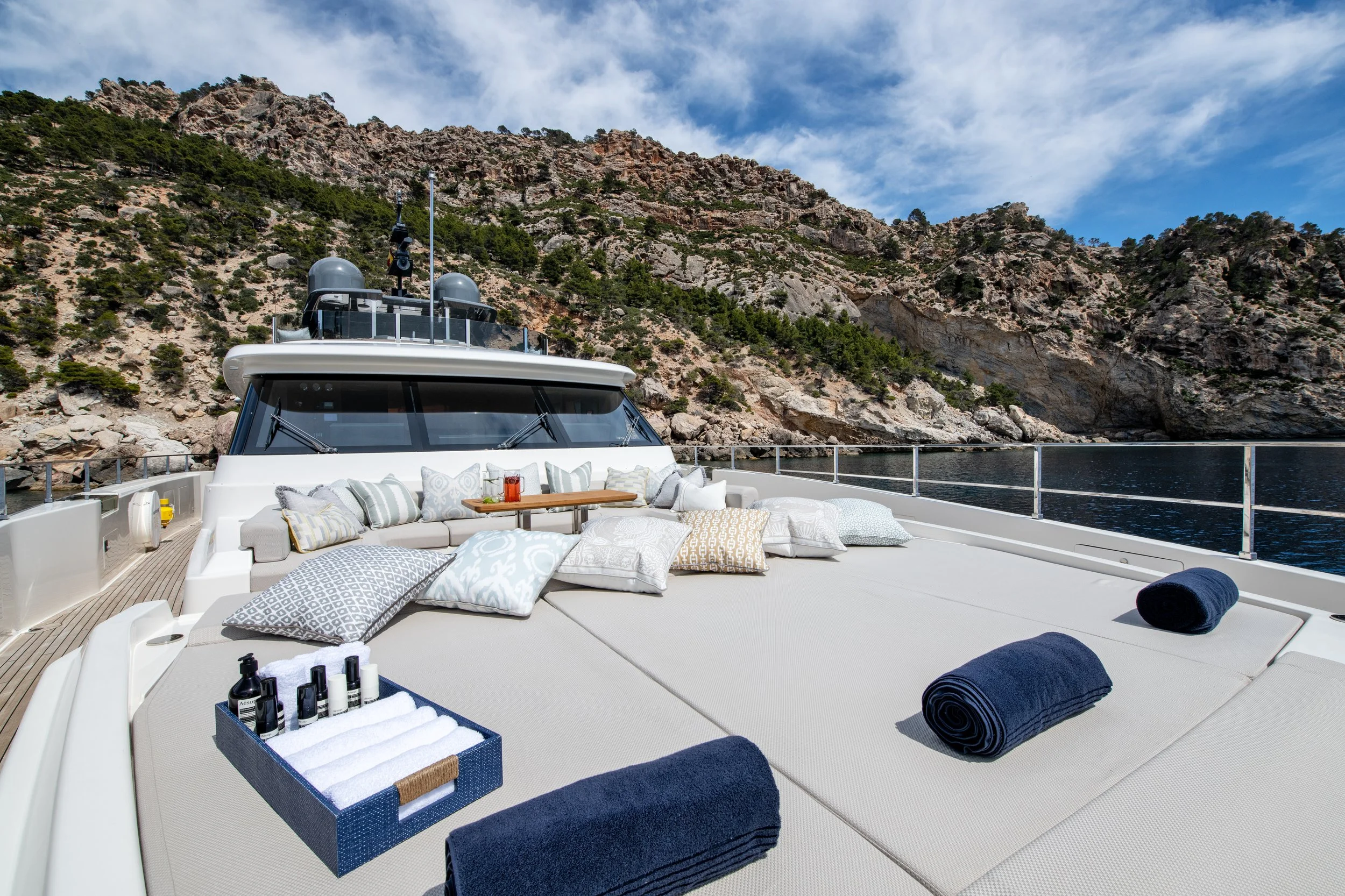 Luxury yacht deck with cushions, rolled towels, and a tray of toiletries, with rocky cliffs and water in the background.