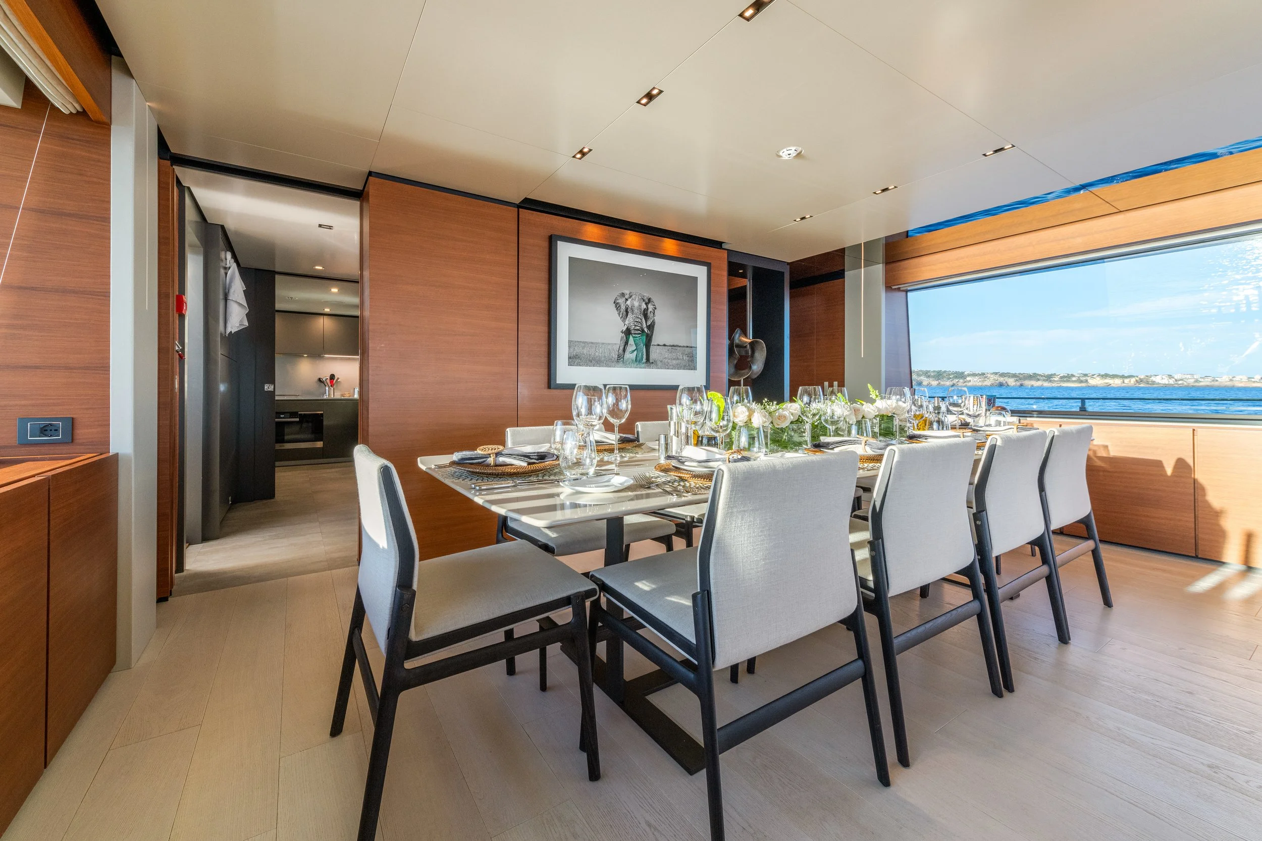 Elegant dining room with a long table set for a meal, featuring white upholstered chairs, glassware, plates, and floral centerpieces. Large windows offer a view of the water and sky outside, with warm wooden walls and ceiling accents.