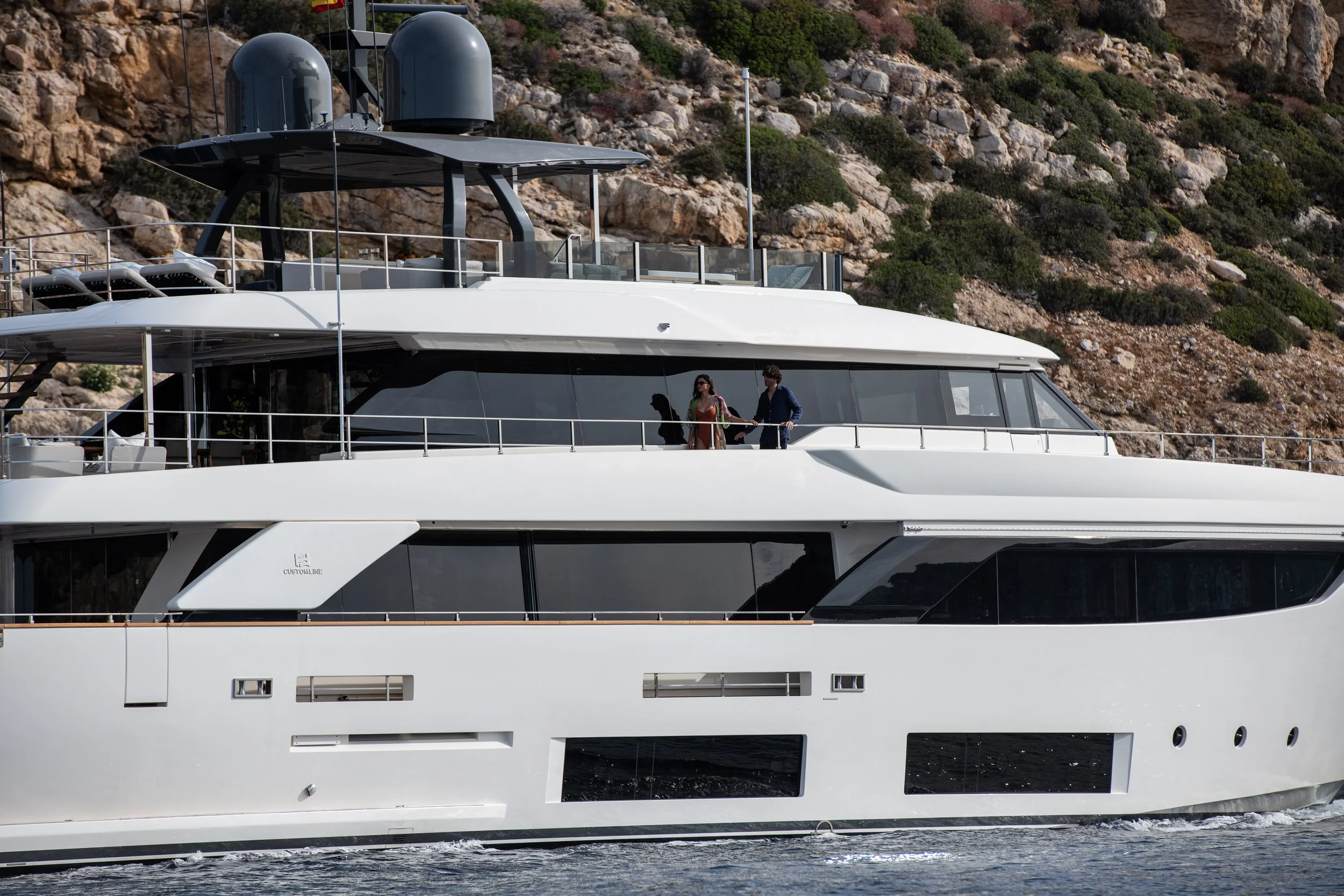 A large luxury white yacht sailing near a rocky shoreline with green bushes. Two people are standing on the upper deck, one in a red dress and the other in a blue suit, holding hands.