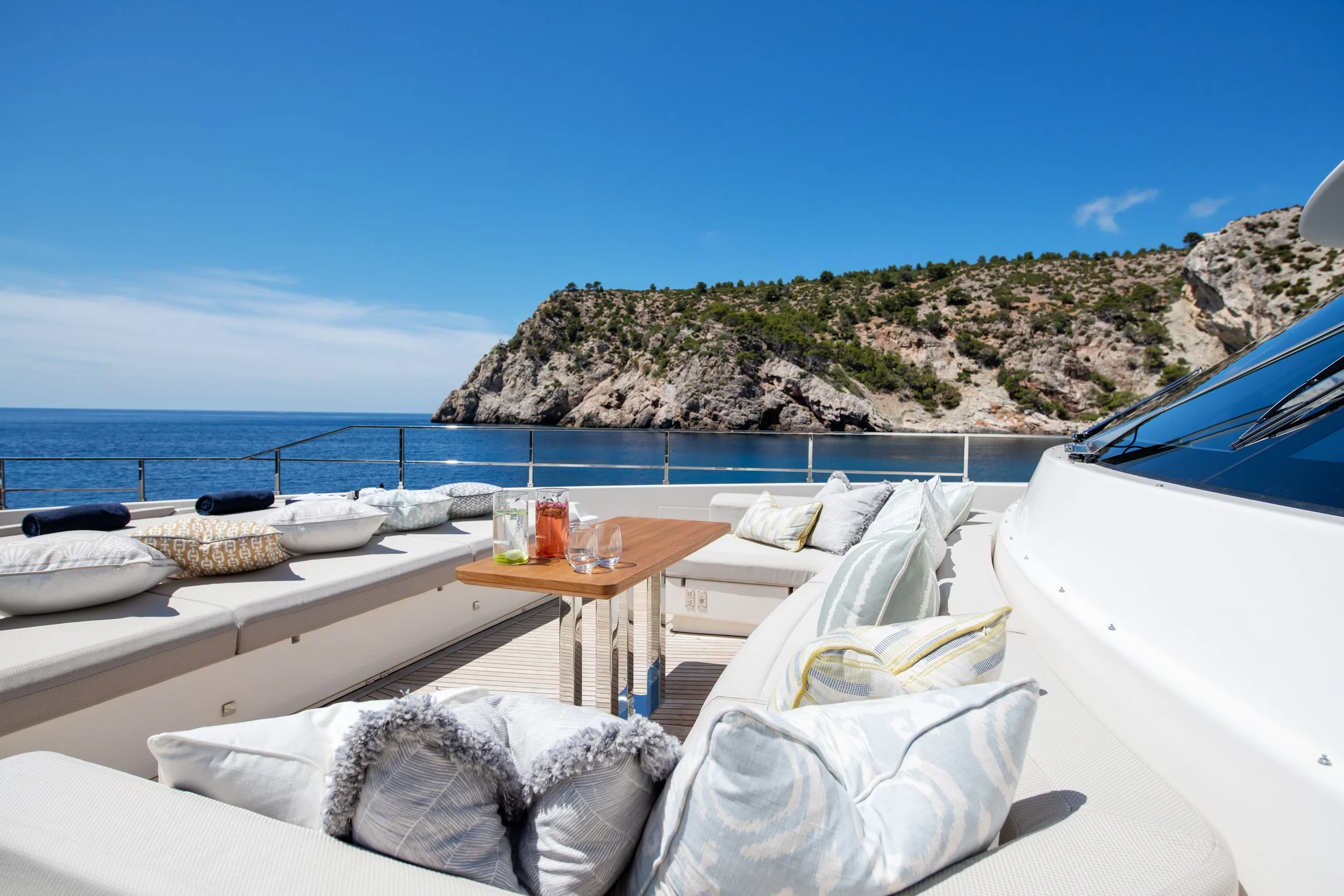 Luxury yacht deck with seating area, pillows, a wooden table with drinks, overlooking the sea and a rocky coastline under a blue sky.