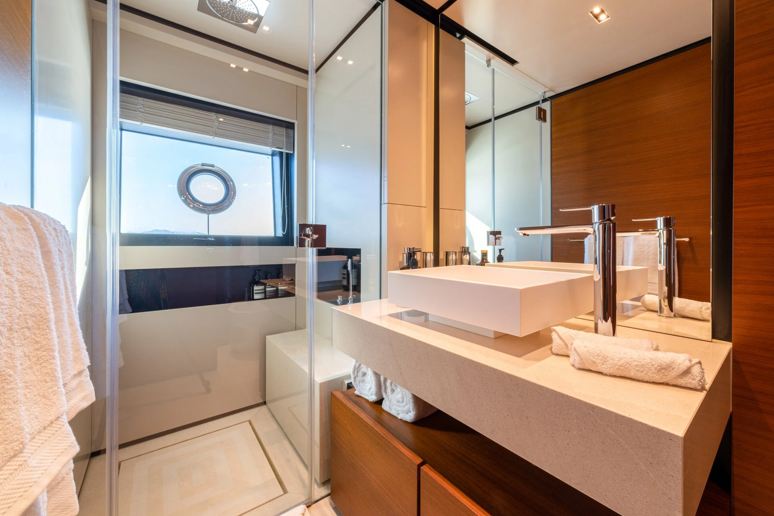 Modern bathroom with a rectangular vessel sink on a beige stone countertop, large mirror, brown wooden cabinet, glass shower enclosure, towels, and a window with a circular feature facing outside.