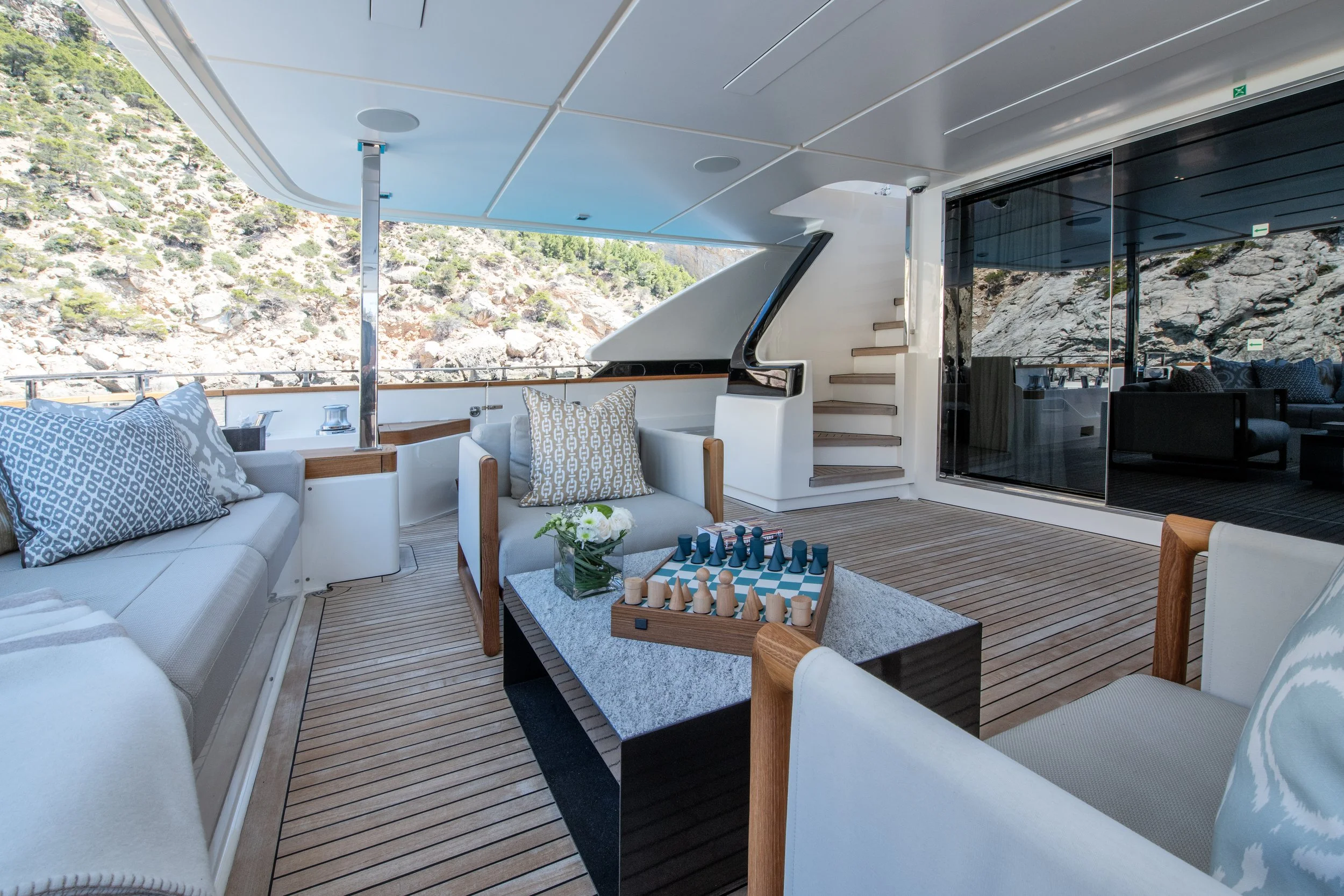 A luxurious yacht deck with white seating, patterned cushions, a chessboard on a table, and a view of rocky terrain through glass sliding doors.