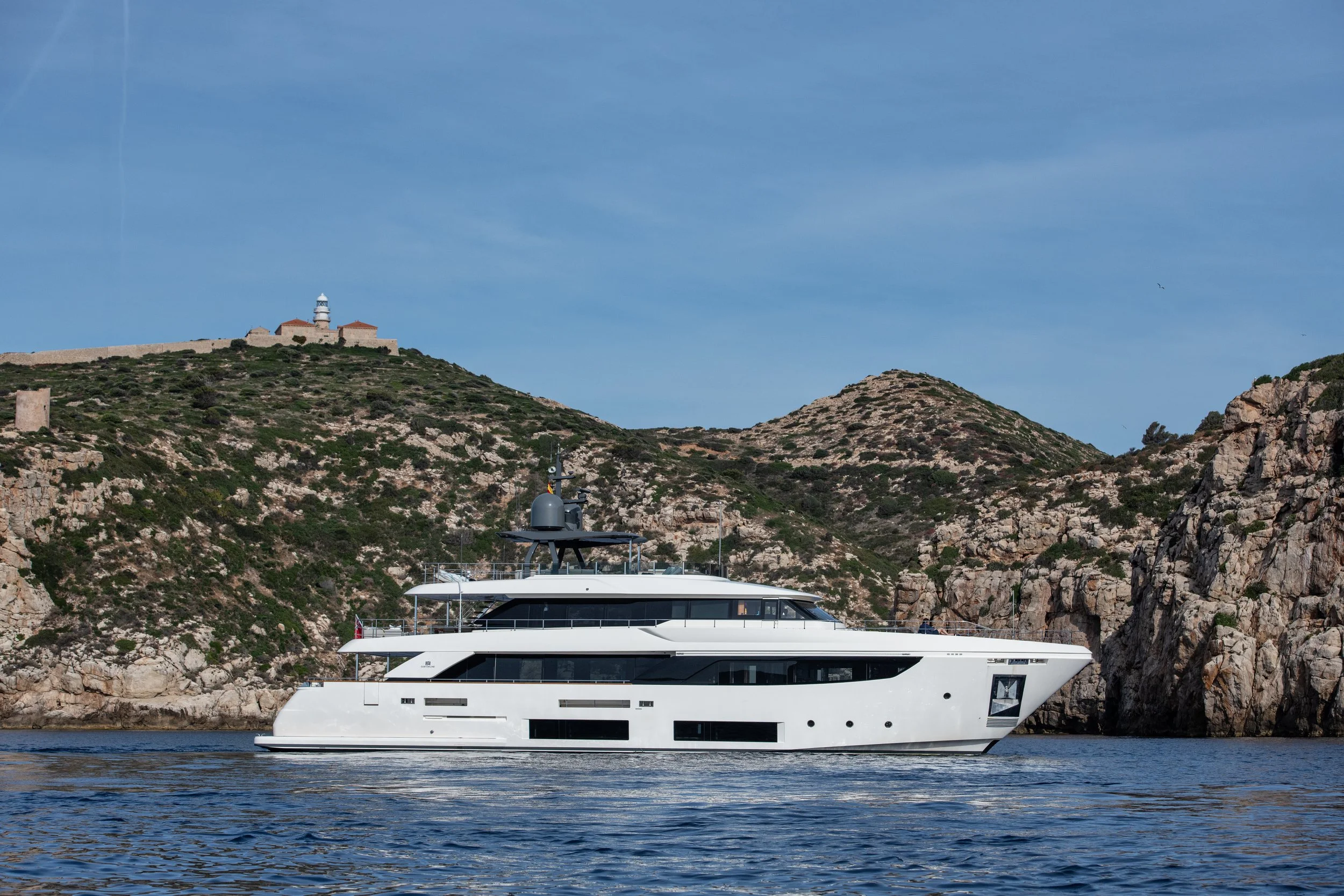 A large white yacht sailing in the water near rocky hills with sparse vegetation, with a lighthouse on a hilltop in the background under a partly cloudy sky.
