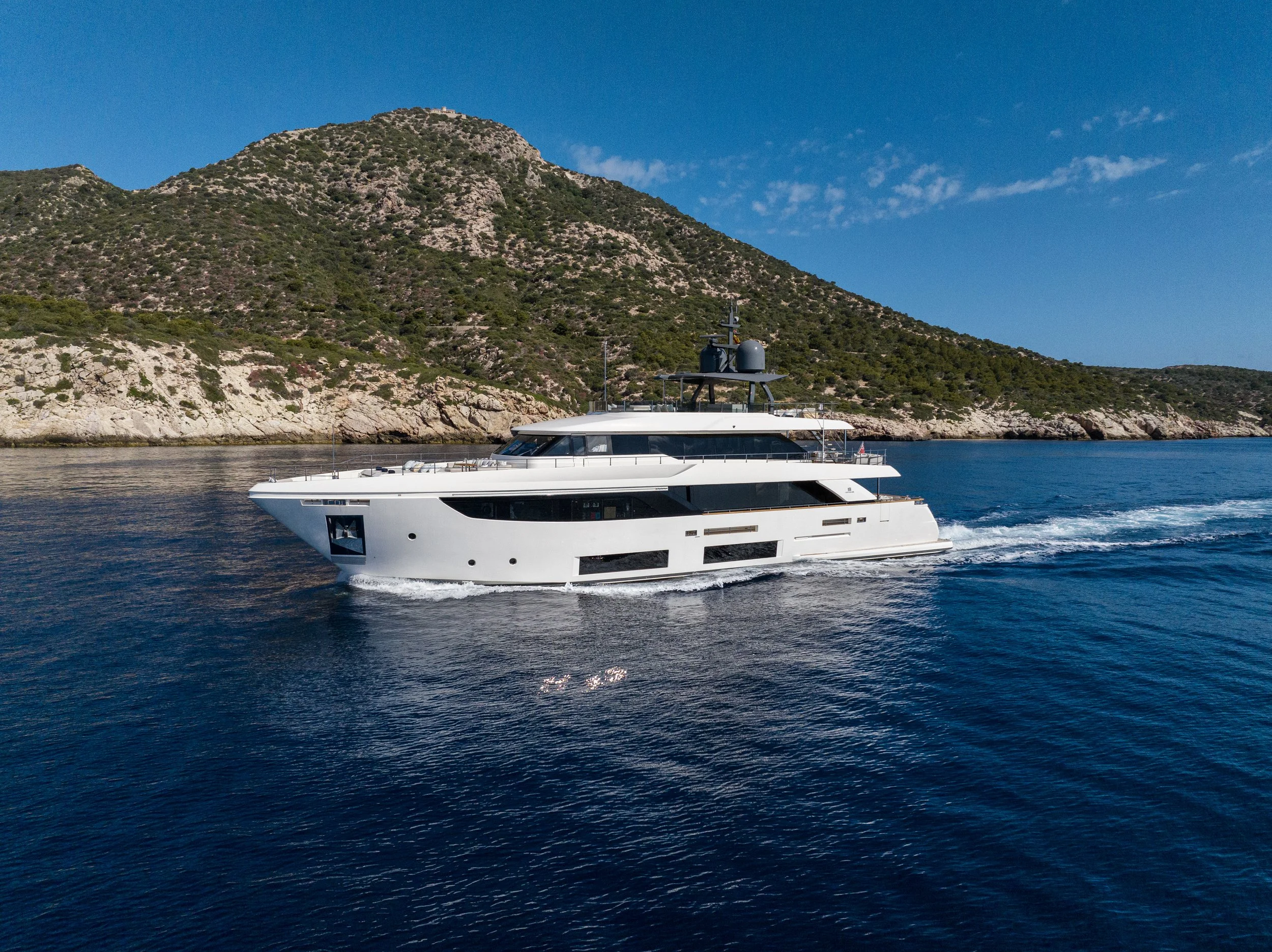 A white yacht sailing near a rocky coastline with a mountain in the background and a clear blue sky.