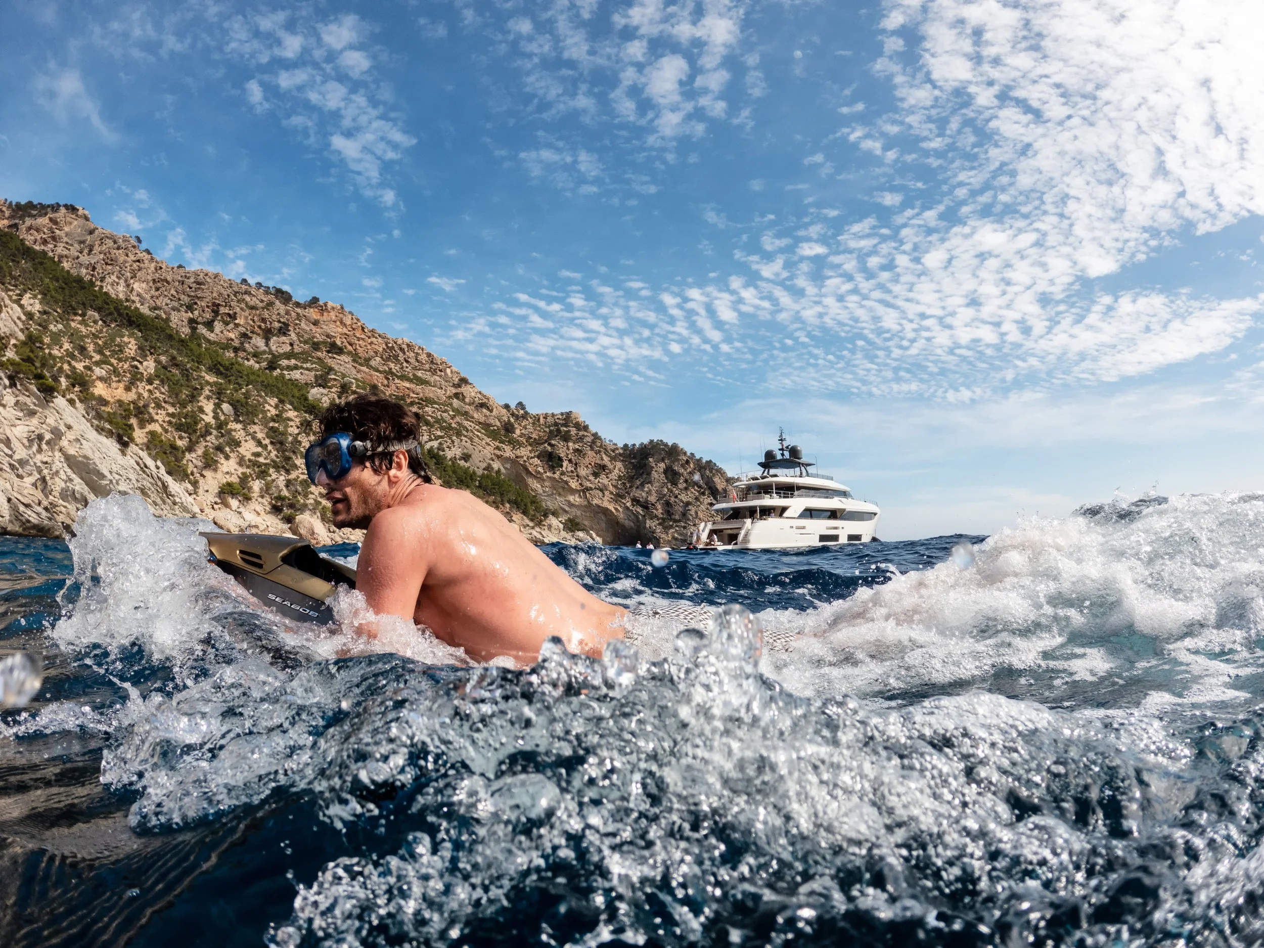 A man swimming in the ocean with a yacht and rocky shoreline in the background under a partly cloudy sky.