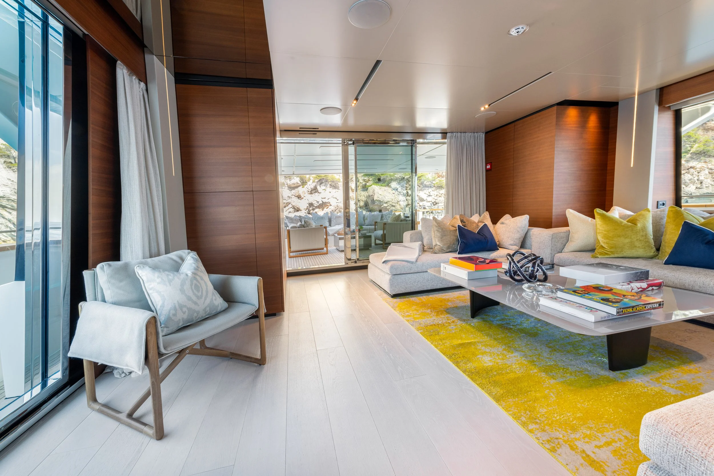 Modern living room with beige sectional sofa, colorful pillows, yellow and blue accents, wooden wall panels, glass sliding doors leading to a balcony with outdoor seating, white armchair near large windows, and a black and white coffee table on a yellow and green rug.