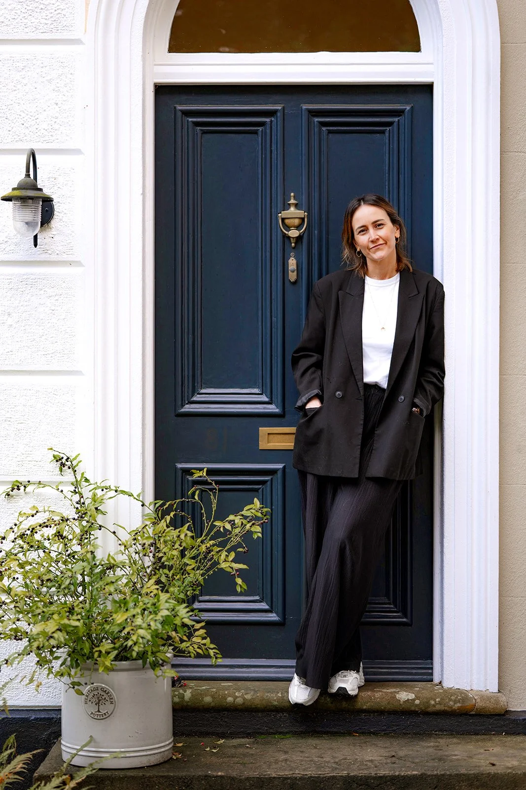 A woman standing casually with her hands in her pockets outside a blue front door of a house, wearing a black blazer, white shirt, black pants, and white sneakers, smiling softly.