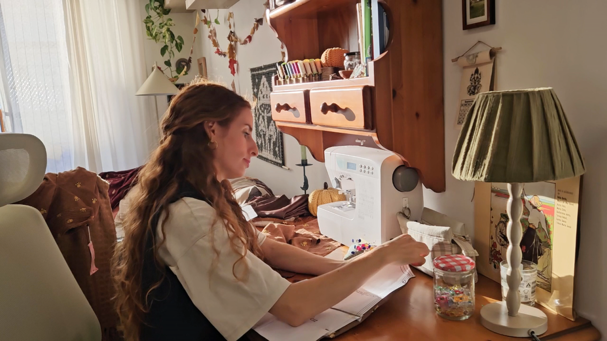 A woman sitting at a wooden desk with a sewing machine, organizing colorful pins and papers, surrounded by autumn-themed decorations and a lamp.