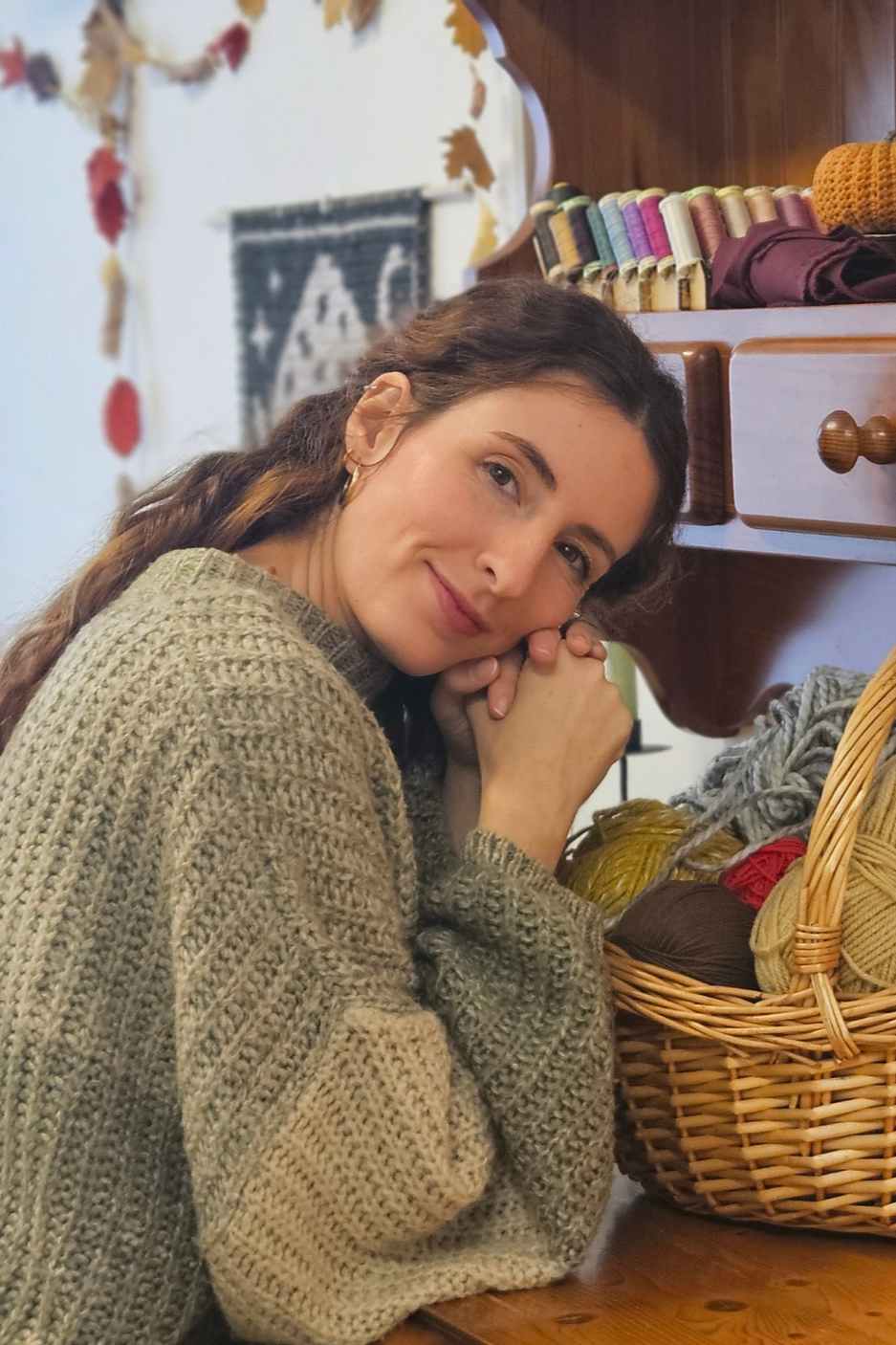 A woman with long brown hair and earrings, wearing a beige knit sweater, sitting at a table with a basket of colorful yarns, smiling and resting her chin on her hands.