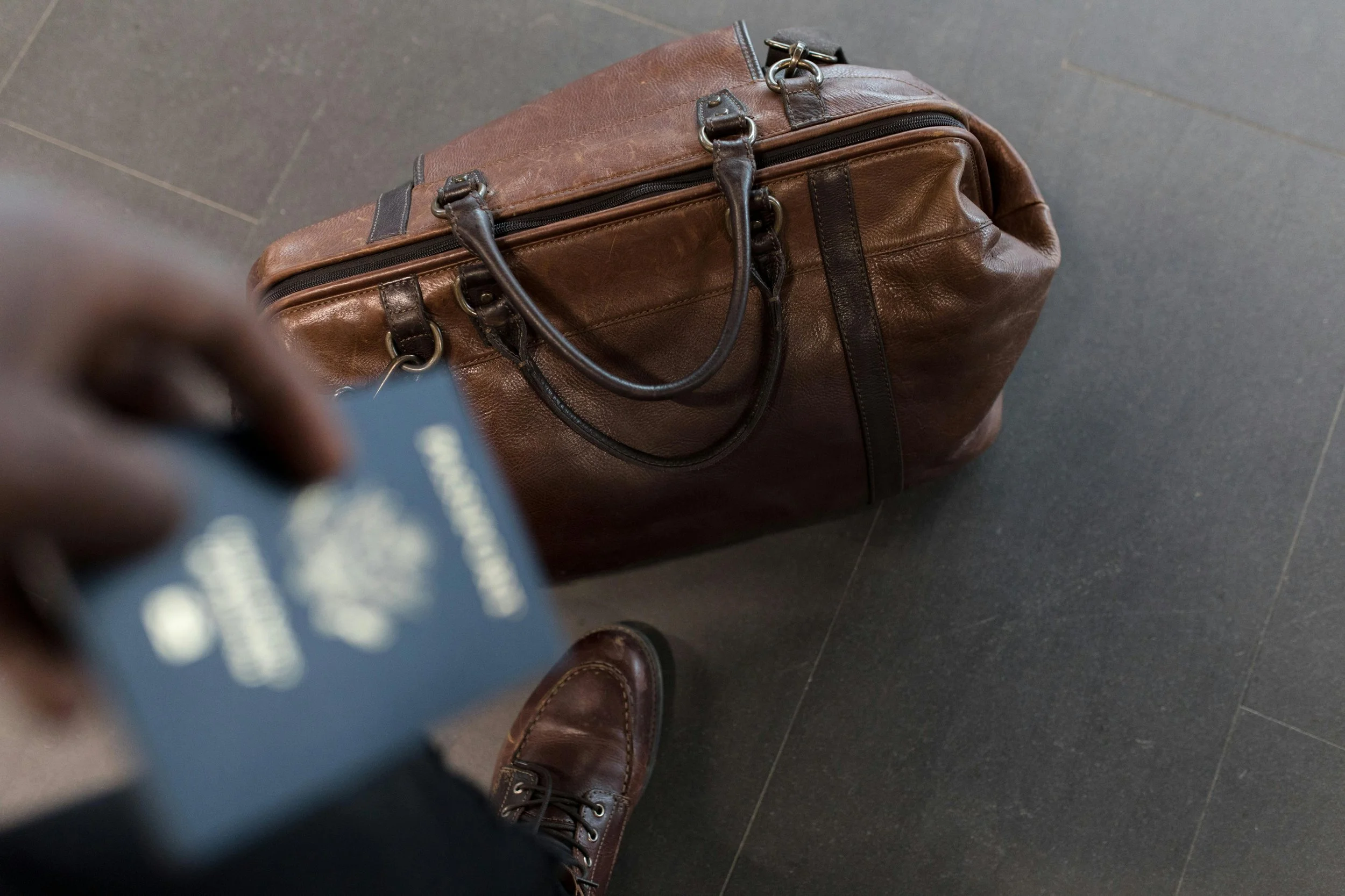 Overhead view of a brown leather travel bag on dark tiled floor, with a person holding a passport and wearing brown leather shoes.