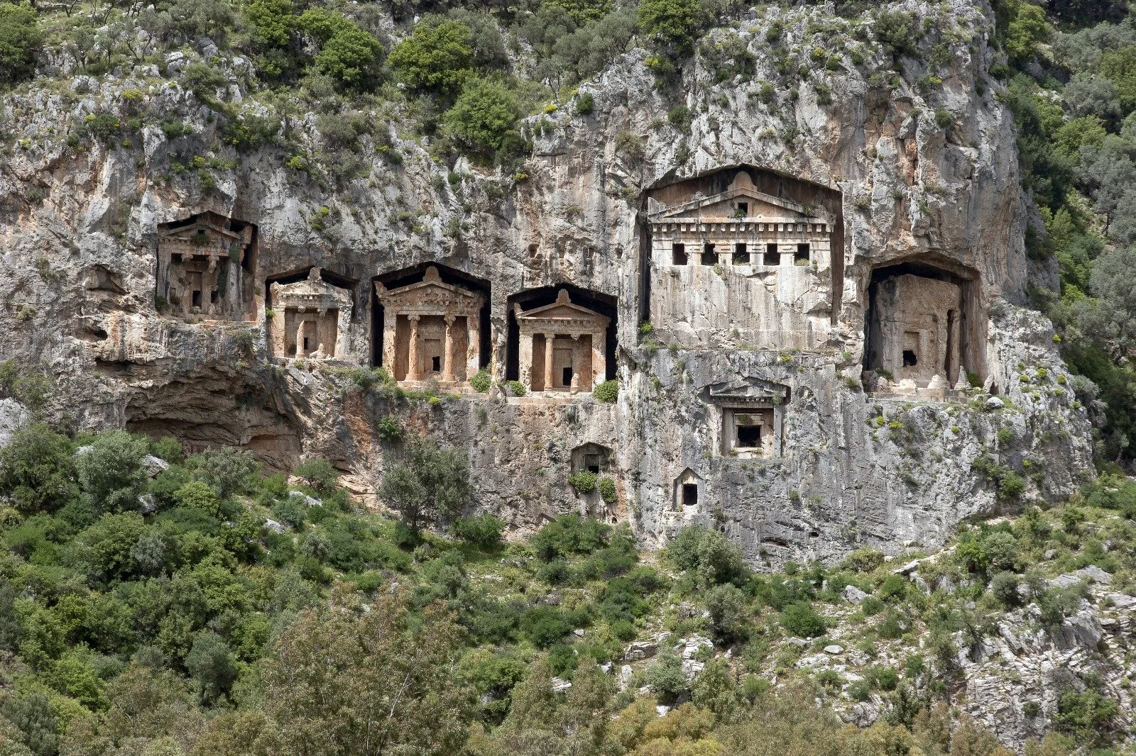 Ancient rock-cut tombs with classical facades carved into a cliff face, surrounded by greenery, at the site of Lycian tombs in Dalyan, Turkey.