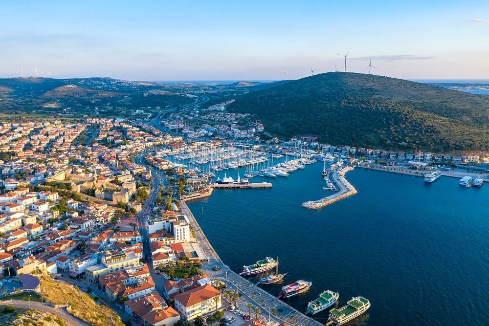 Aerial view of a coastal city with a marina, boats, houses, and a hill with wind turbines.