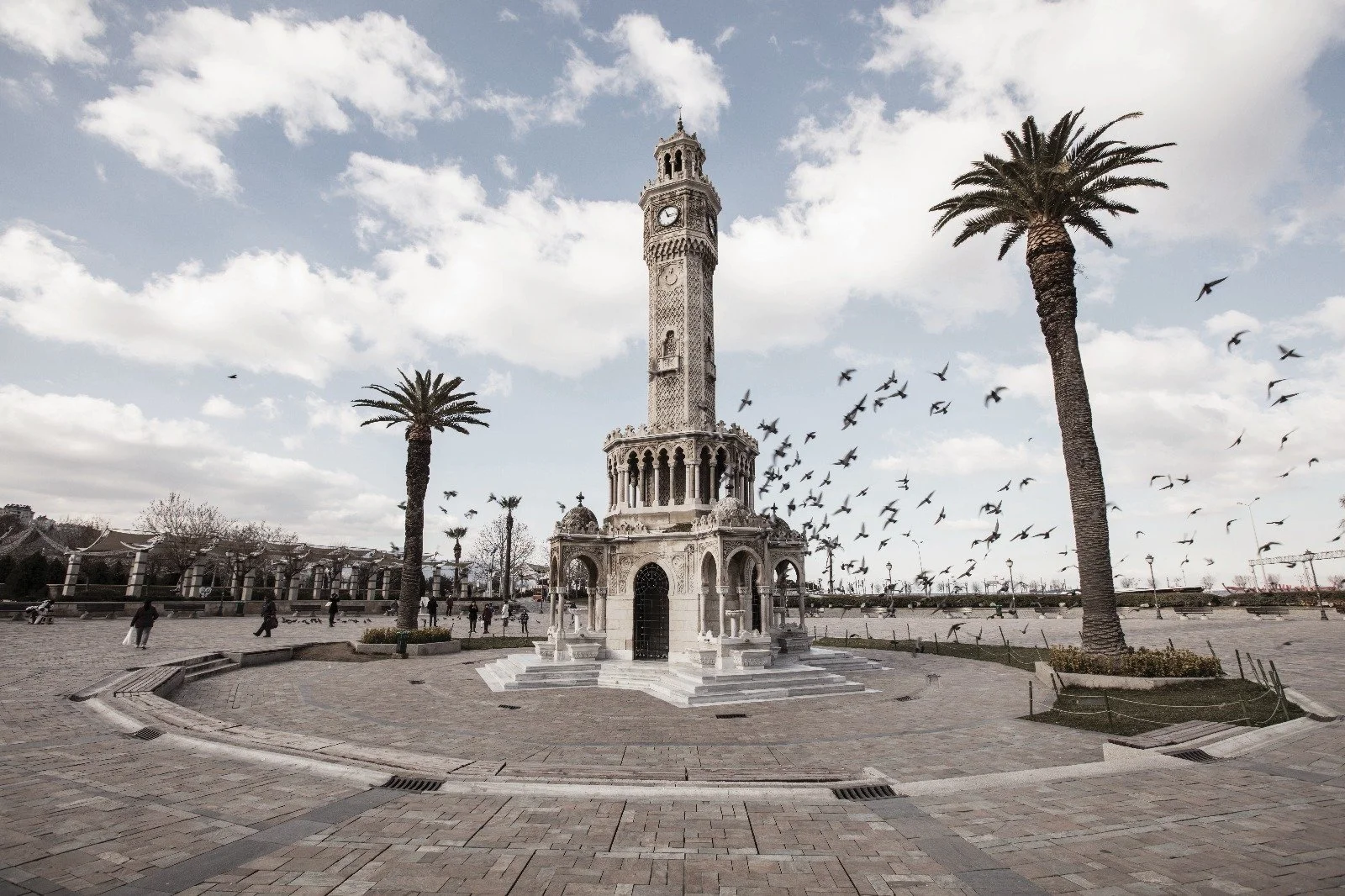 A tall ornate clock tower with decorative arches and three domes on a paved plaza, flanked by two tall palm trees, with birds flying in a cloudy sky.