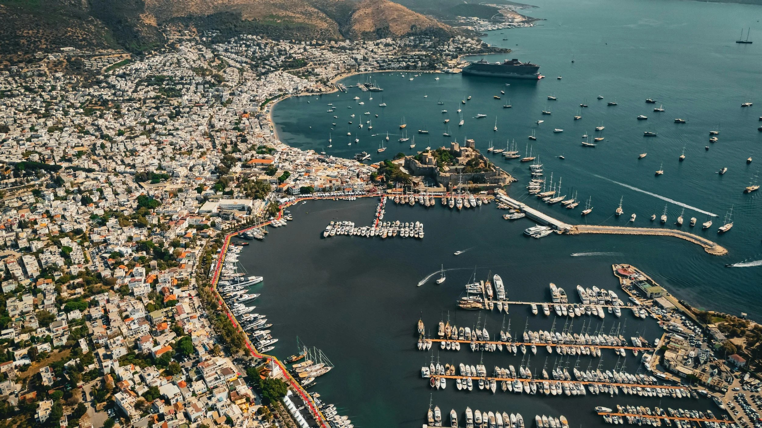 Aerial view of a coastal city with numerous boats and yachts docked in marinas, surrounding a hilly landscape and waterfront buildings.