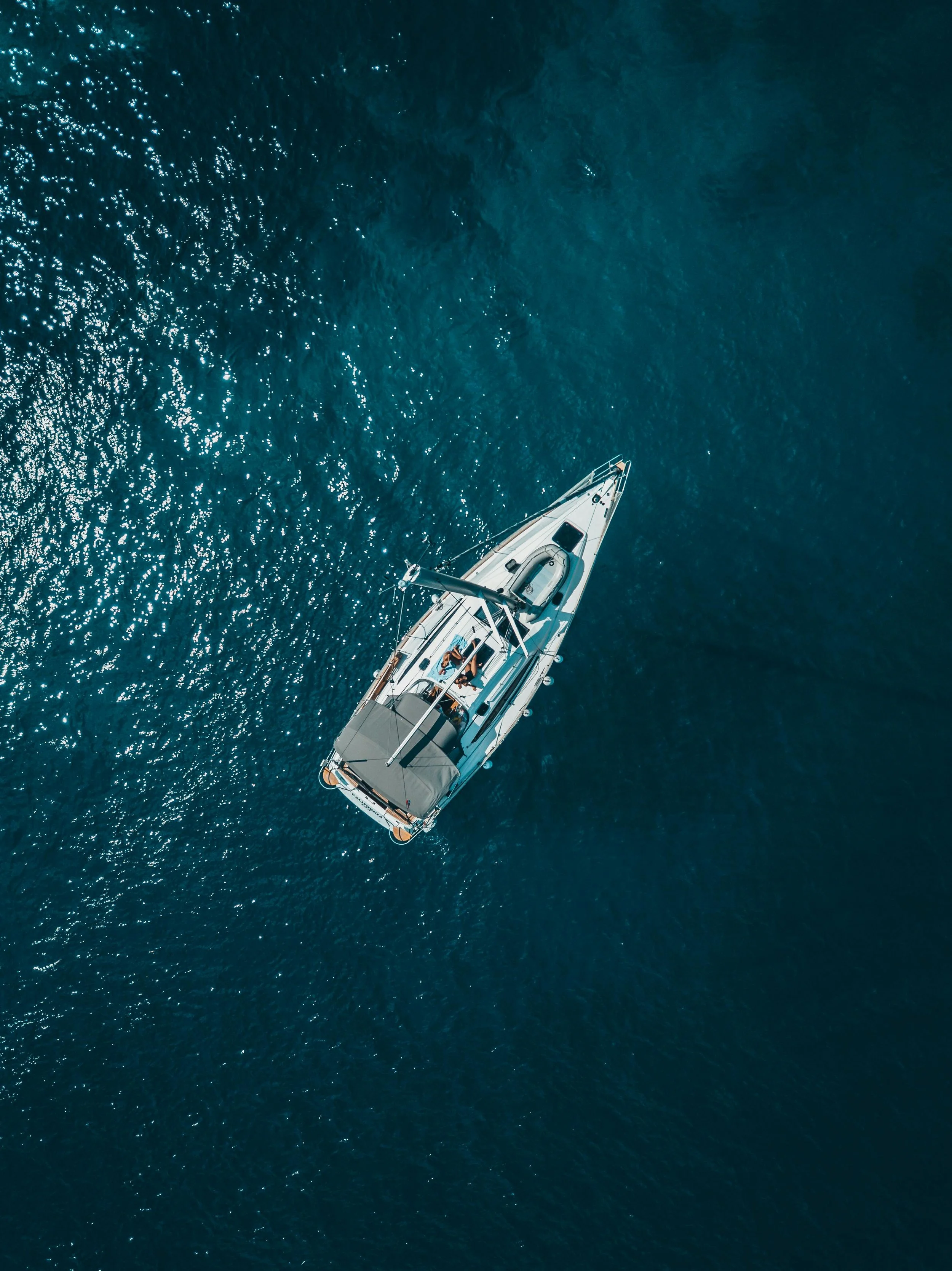 An aerial view of a sailboat on the open water with the sun reflecting off the surface.