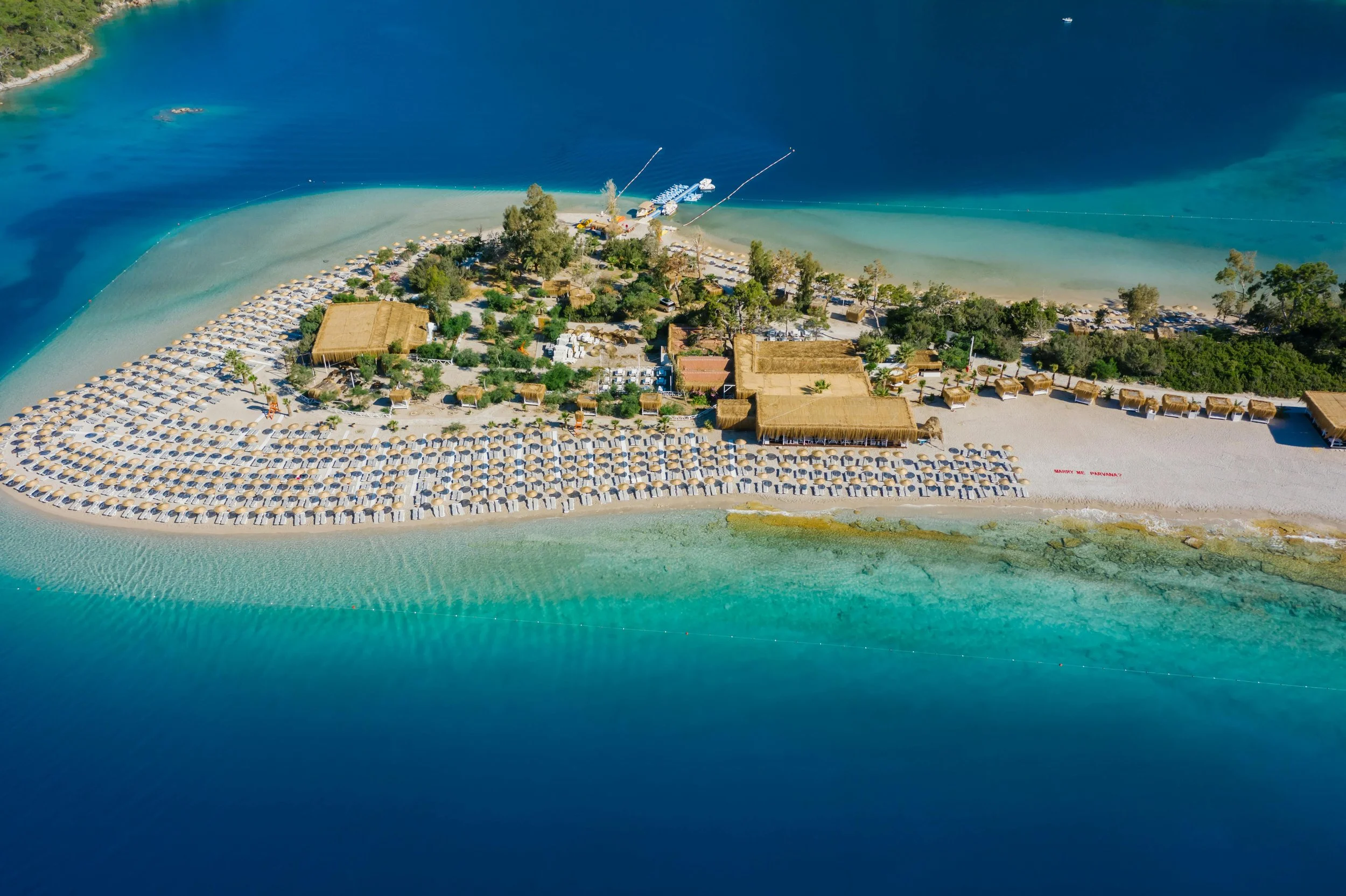 Aerial view of a beach with many umbrellas and sun loungers, surrounded by water and greenery.