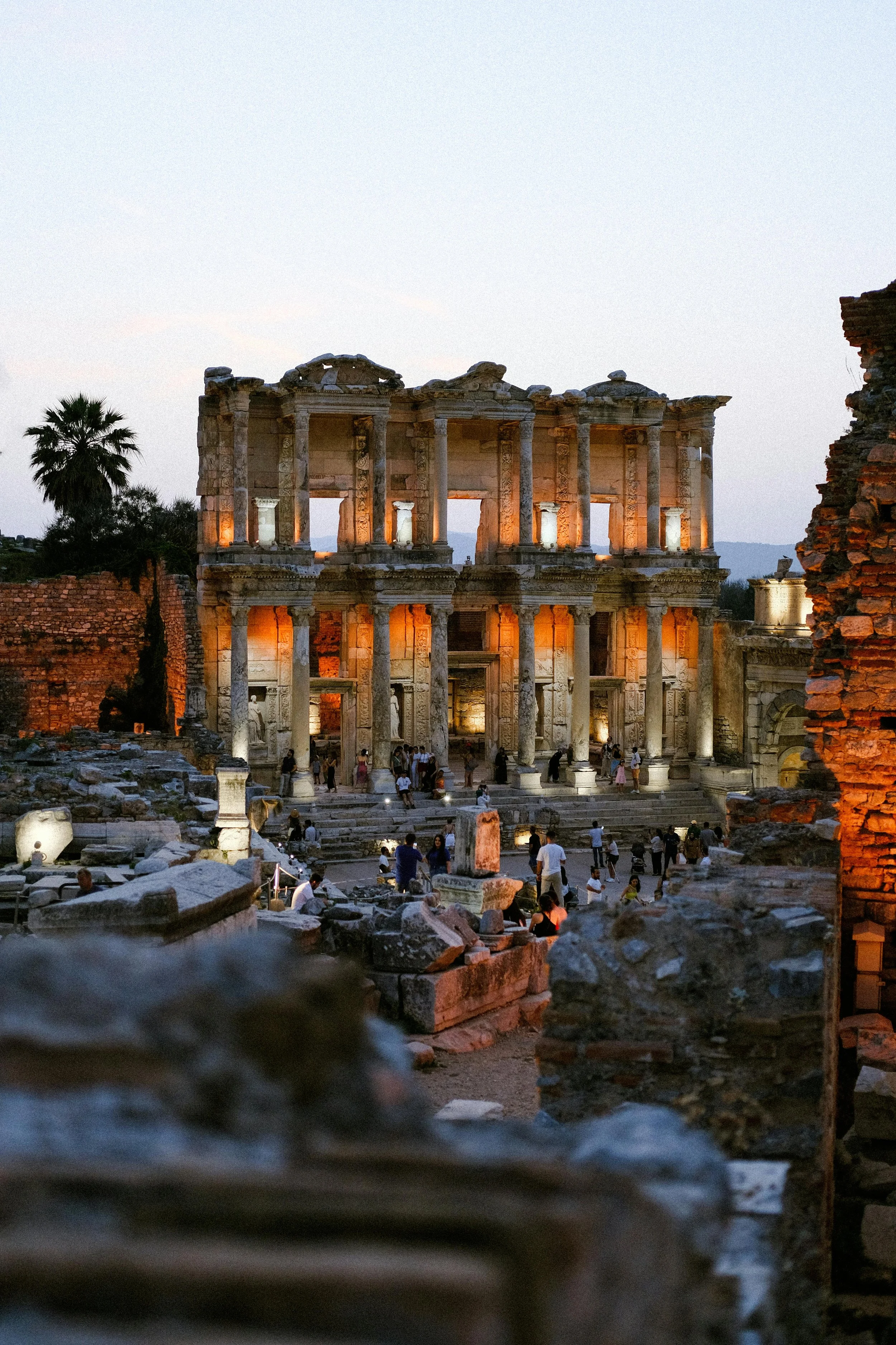 Ancient Roman ruins at dusk, with illuminated two-story structure featuring columns and arches, scattered stones, and visitors exploring the site.