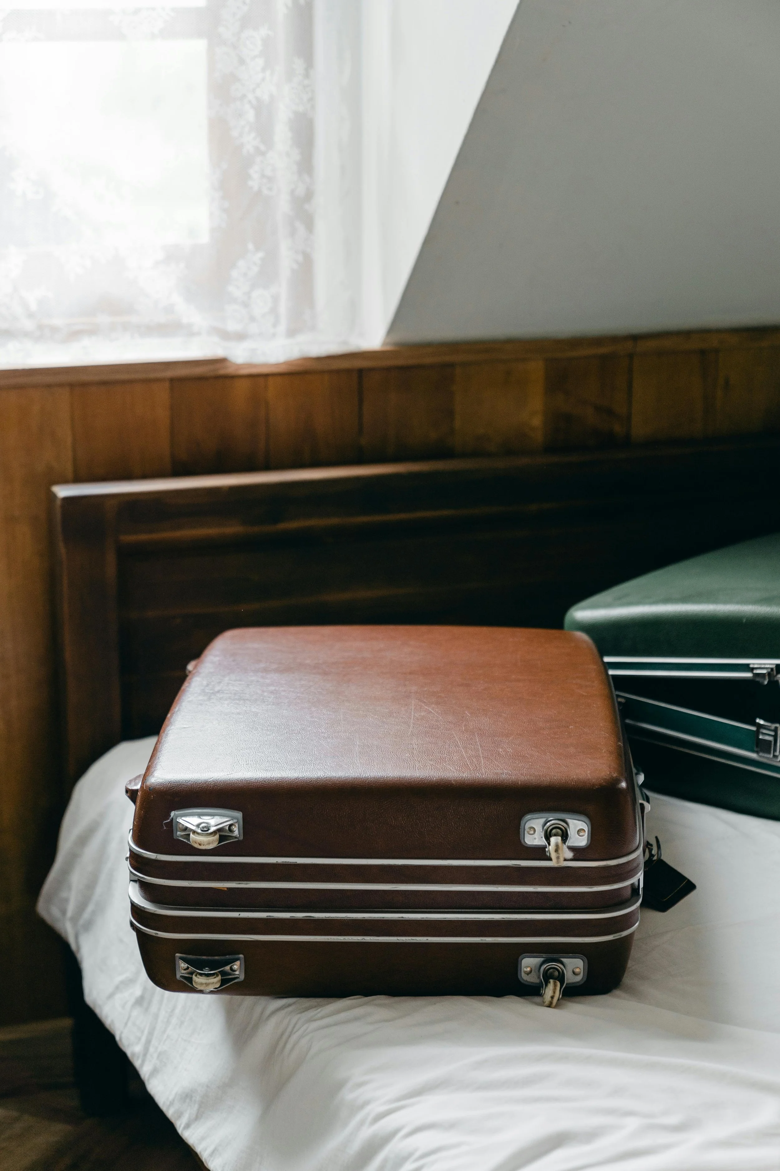 A vintage brown hard-shell suitcase with metal latches placed on a bed with a white sheet, beside a green suitcase, in a room with wooden walls and lace curtains.