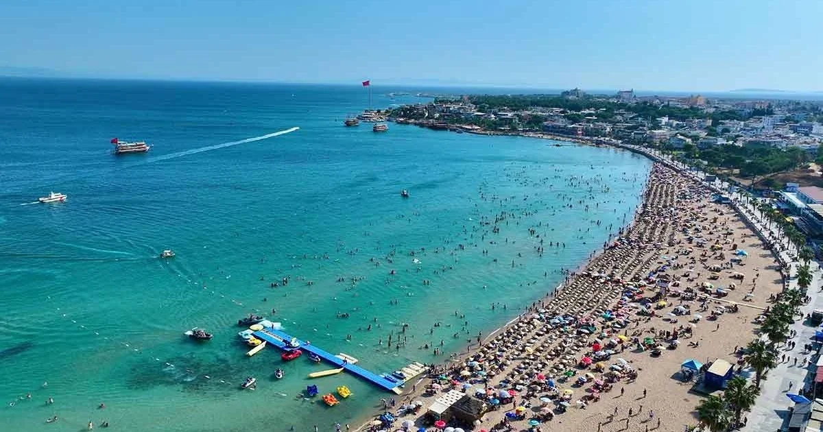 A crowded beach with many umbrellas and people, boats in the water, and a cityscape in the background.