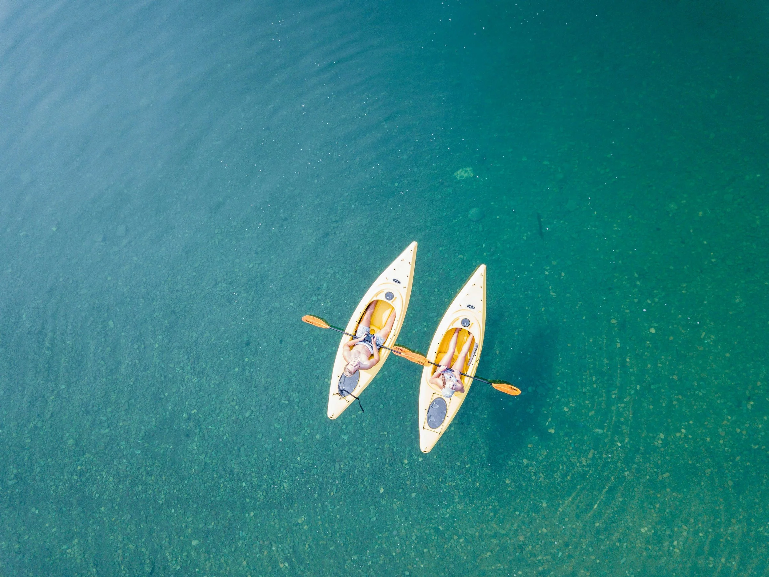 Two people kayaking on clear blue water from an aerial view.