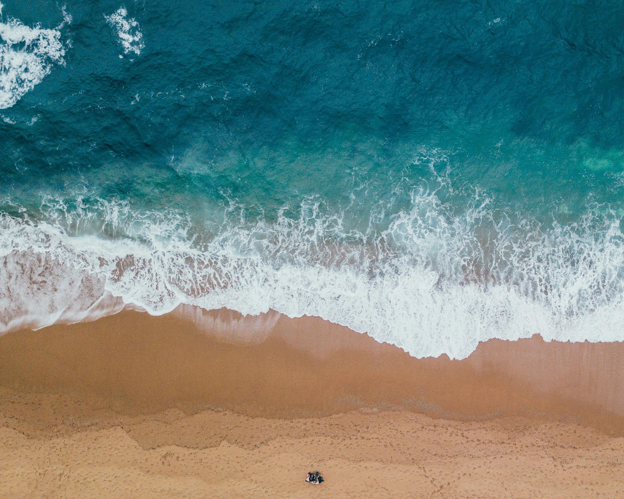 Aerial view of ocean waves crashing onto sandy beach with small boat visible on the sand.
