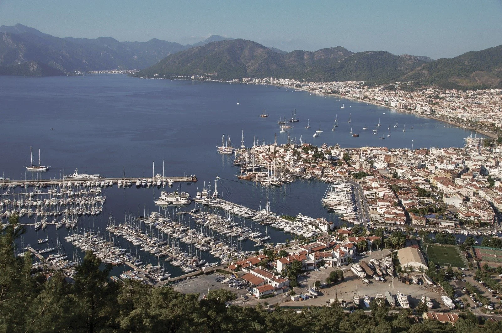 Aerial view of a marina with numerous sailboats and yachts docked, overlooking a coastal city with a bay surrounded by mountains.