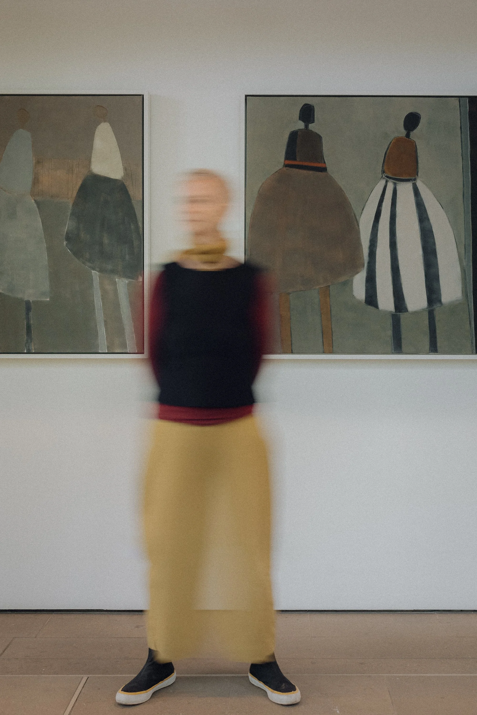 Person with short grey hair and mustard trousers, wearing black and white boots, walking past two large abstract paintings of figures in dresses, in an art gallery.