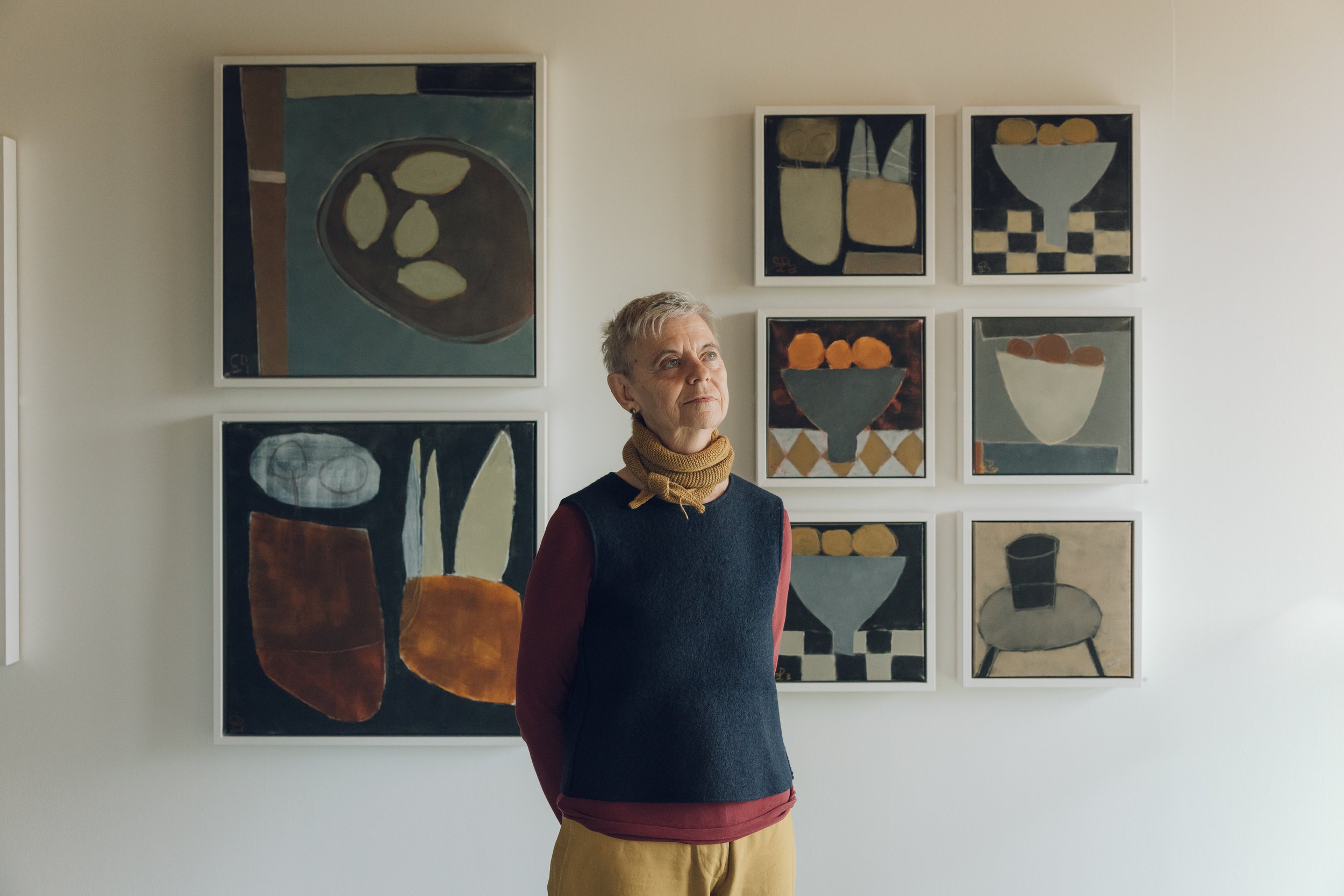 A woman with short grey hair standing in front of a wall with six modern abstract paintings, wearing a black top, a tan scarf, and mustard trousers.