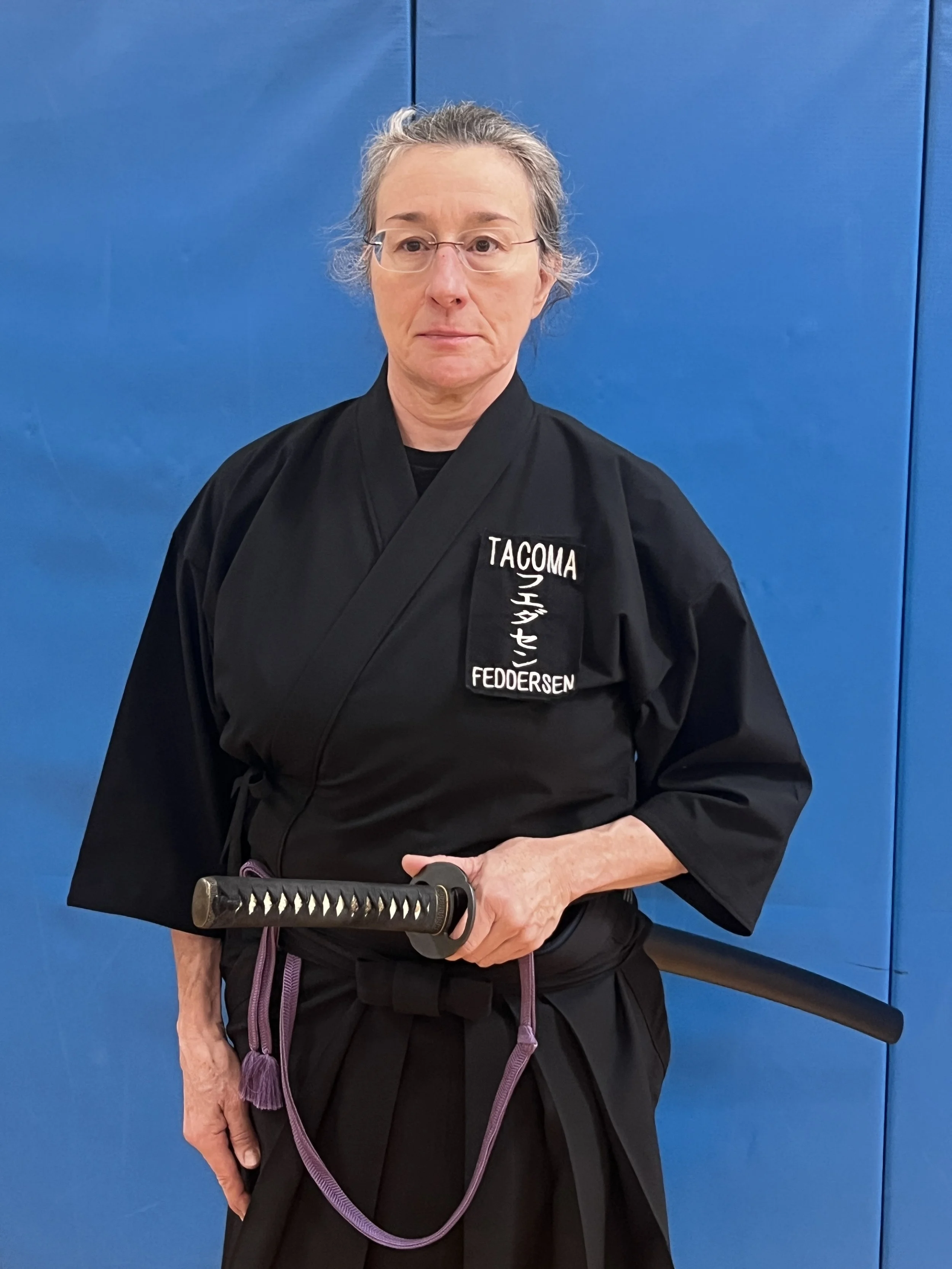 A Iaidoka Sensei in a black martial arts uniform holding a katana sword, standing in front of a blue wall.