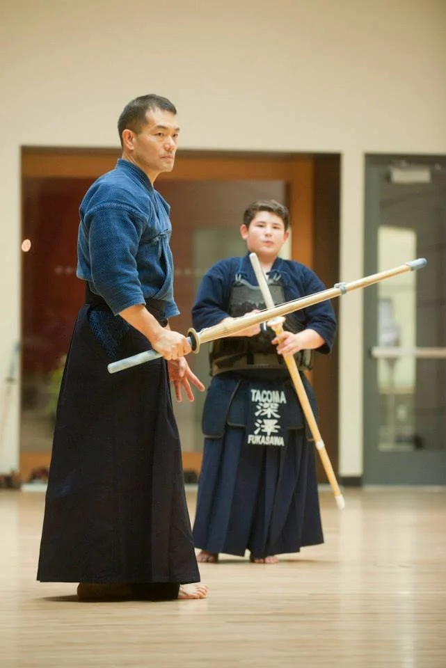 Two martial artists practicing Aikido in a dojo, wearing traditional uniforms and holding wooden practice swords.