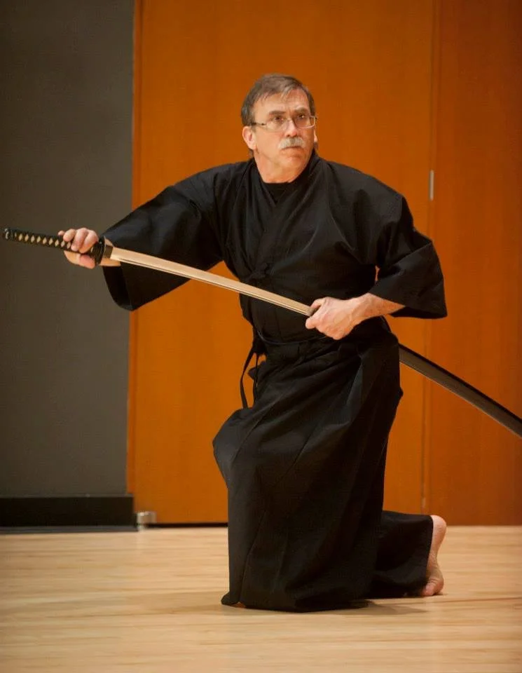 A man wearing a black martial arts uniform practicing with a katana sword indoors on a wooden floor.