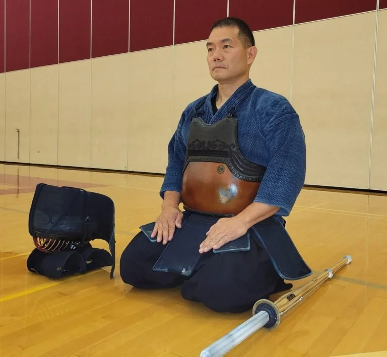 A Kendoka Sensei in traditional attire kneeling on the gym floor, wearing protective gear and surrounded by training equipment.