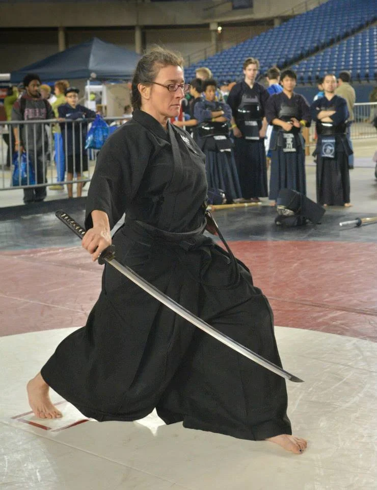 A woman in traditional martial arts attire practicing with a sword during a martial arts demonstration or competition, with several onlookers watching.