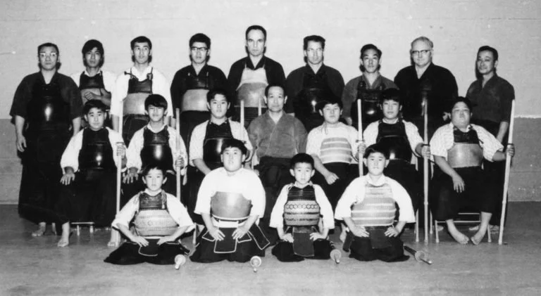 A group of young kendo practitioners, wearing traditional uniforms, sitting and standing with their instructors in a dojo, black and white photo.