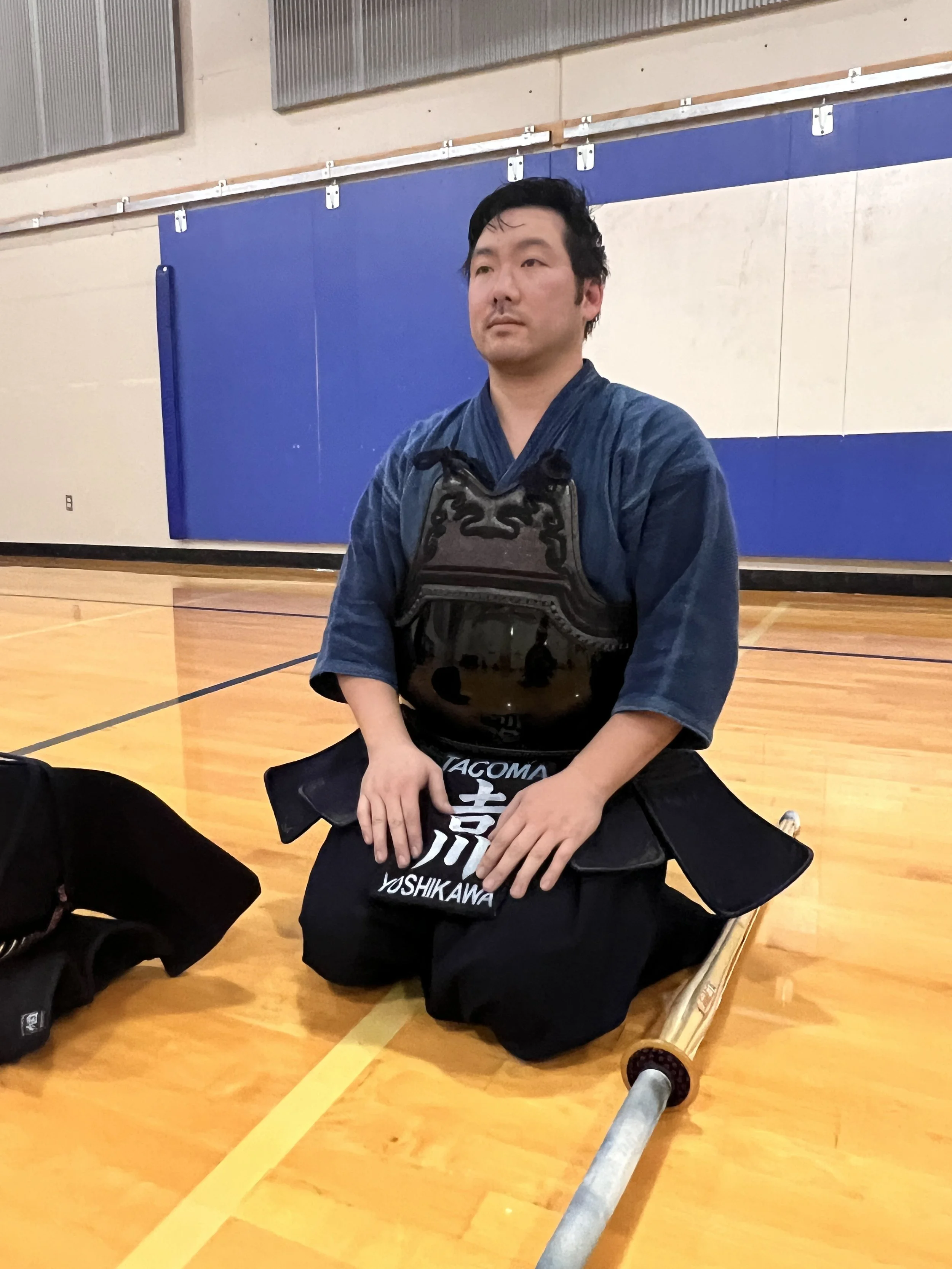 A Kendoka Sensei dressed in traditional Kendo armor, sitting on the gym floor with a Kendo sword resting on the ground beside him.