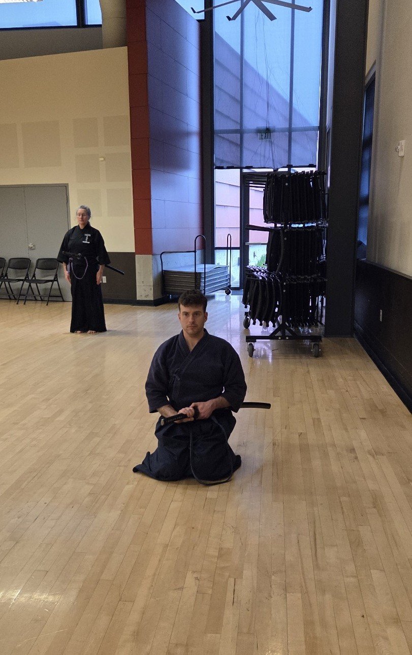 Two martial artists practicing kendo drills in a spacious indoor room. One man kneeling with a shinai, the other standing in the background with a shinai hanging from his side.