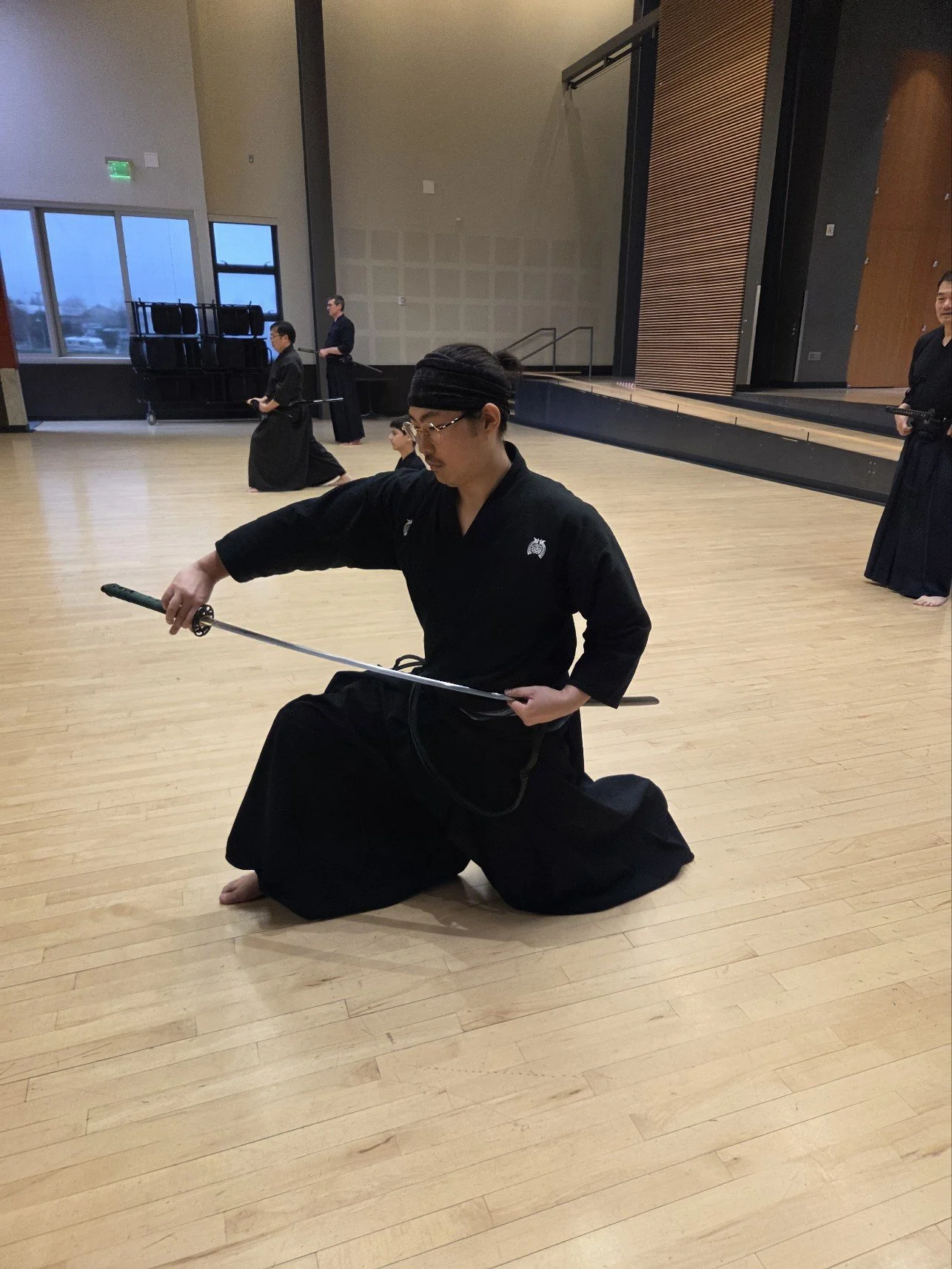 Person practicing martial arts with a sword in a gym or dojo, kneeling on one knee, dressed in traditional black attire.