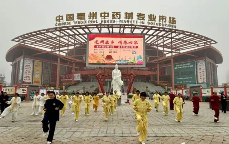 People in yellow and red traditional Chinese clothing running in front of the Chinese Medicinal Herbs Market in Bozhou, China, with a statue and a large digital screen in the background.