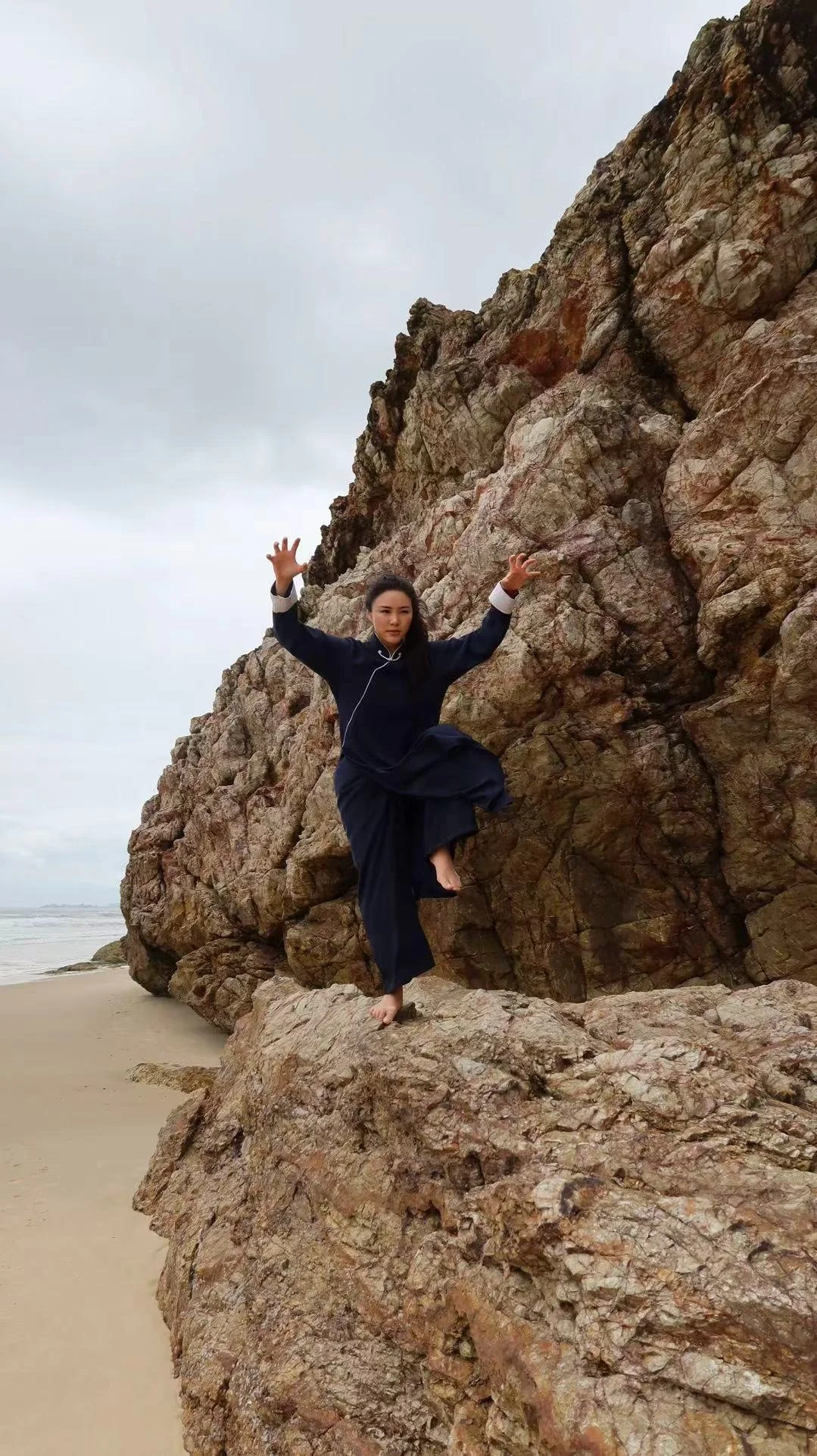A woman in dark clothing balancing on a rock formation at the beach with an overcast sky in the background.