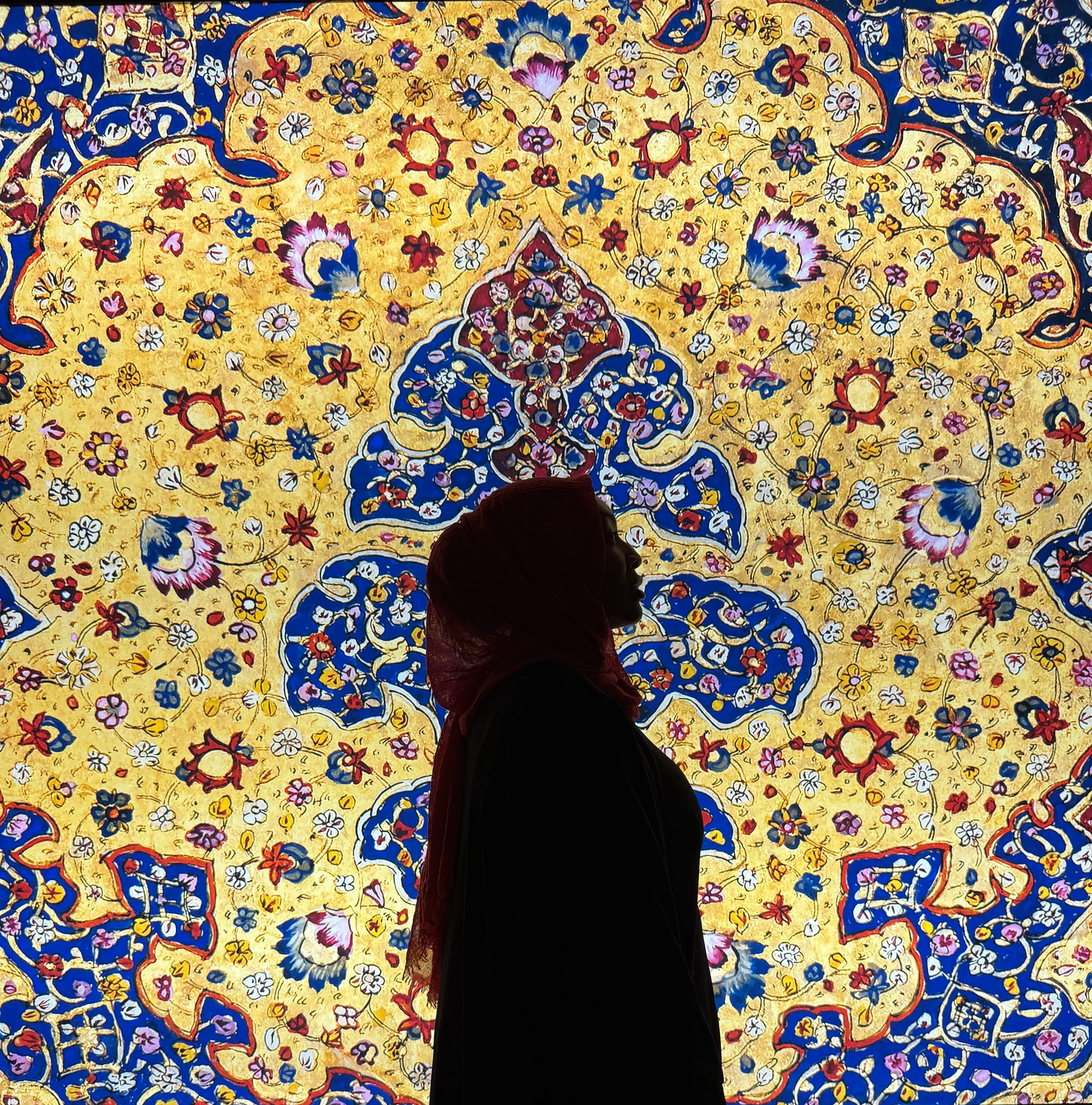 Silhouette of a woman wearing a headscarf in front of a vibrant, ornate, and colorful tapestry or mural with intricate floral and geometric patterns.