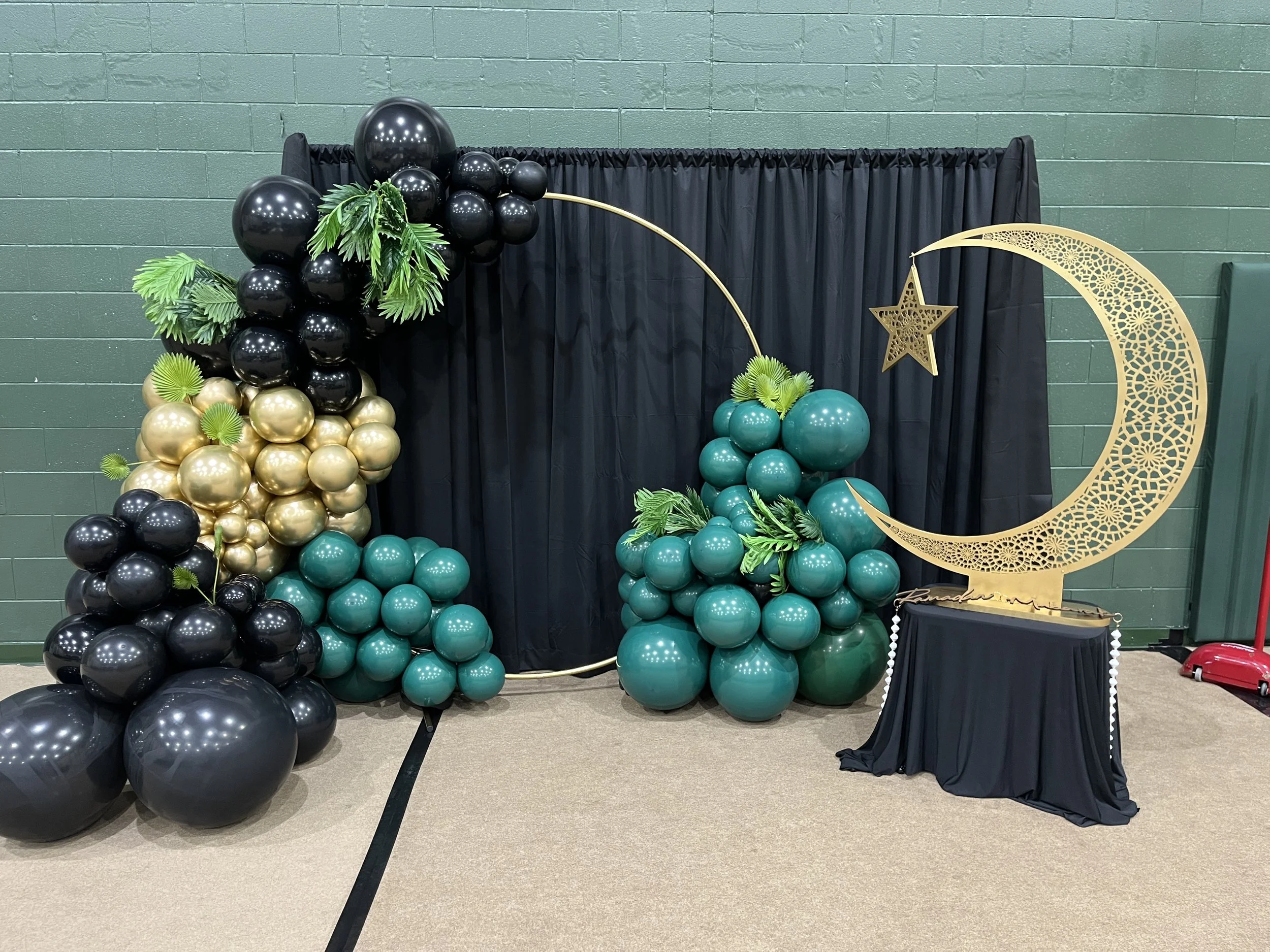 Decorative setup with black, gold, and teal balloons, a black curtain backdrop, and a gold crescent moon and star stand.