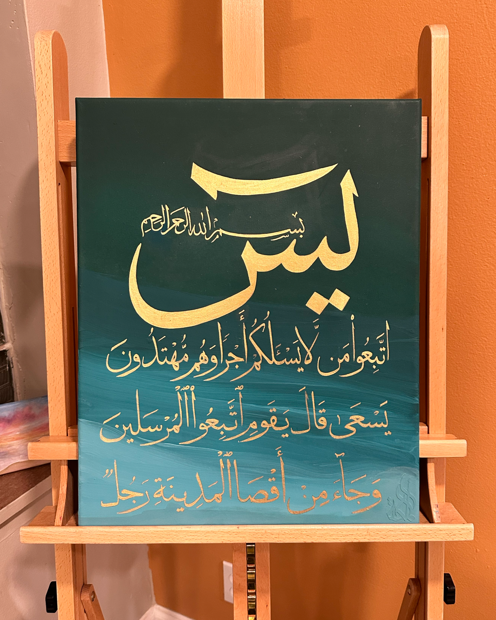 A calligraphic artwork of Islamic text in Arabic on a gradient blue background, mounted on a wooden easel.