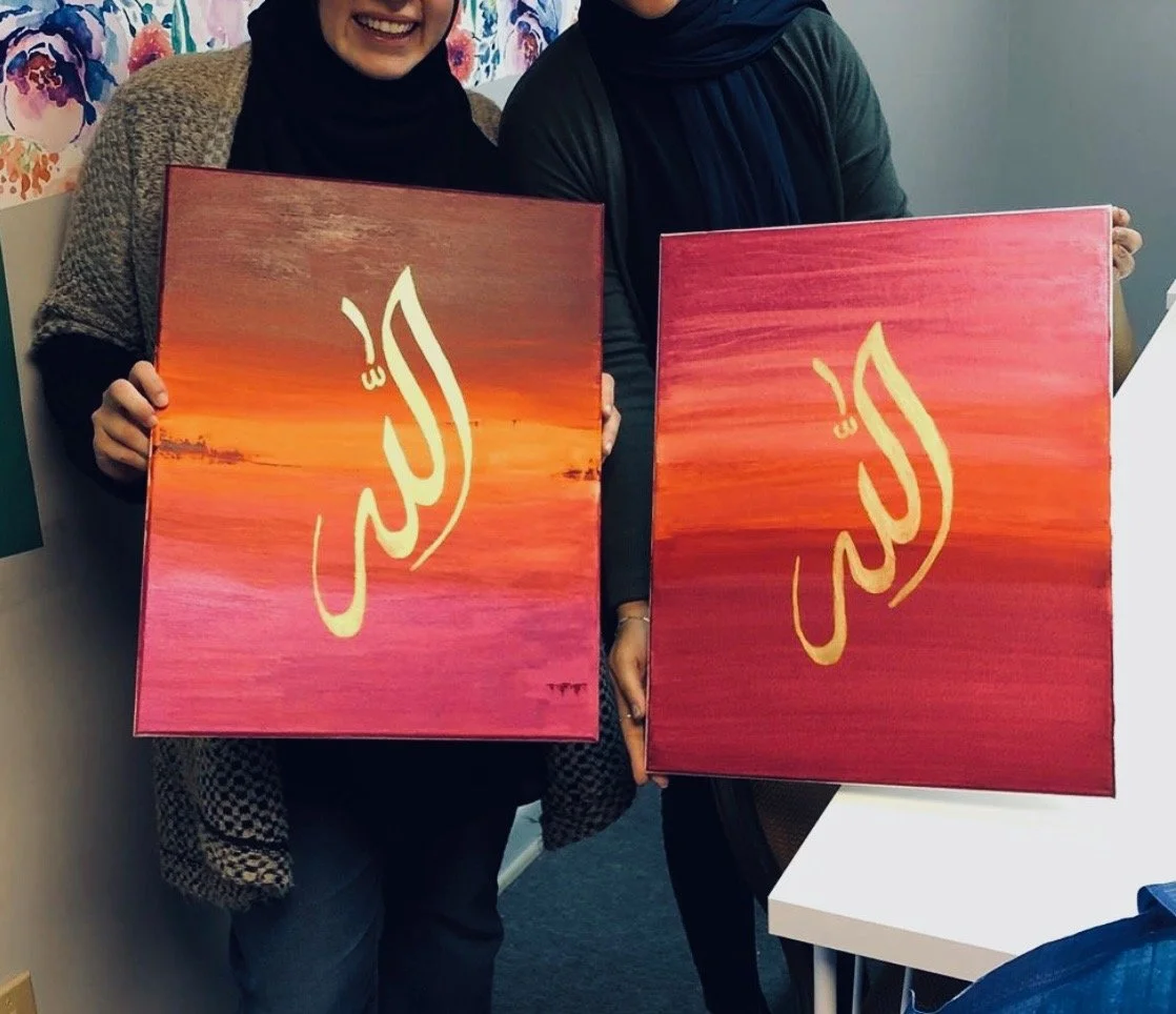 Two women holding square paintings with sunset scenes and Arabic calligraphy in gold.