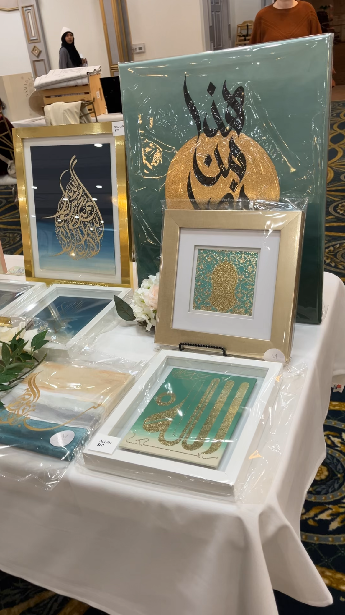 Display of Islamic calligraphy artwork on a table, including framed pieces with gold and blue designs, featuring phrases in Arabic with decorative elements.