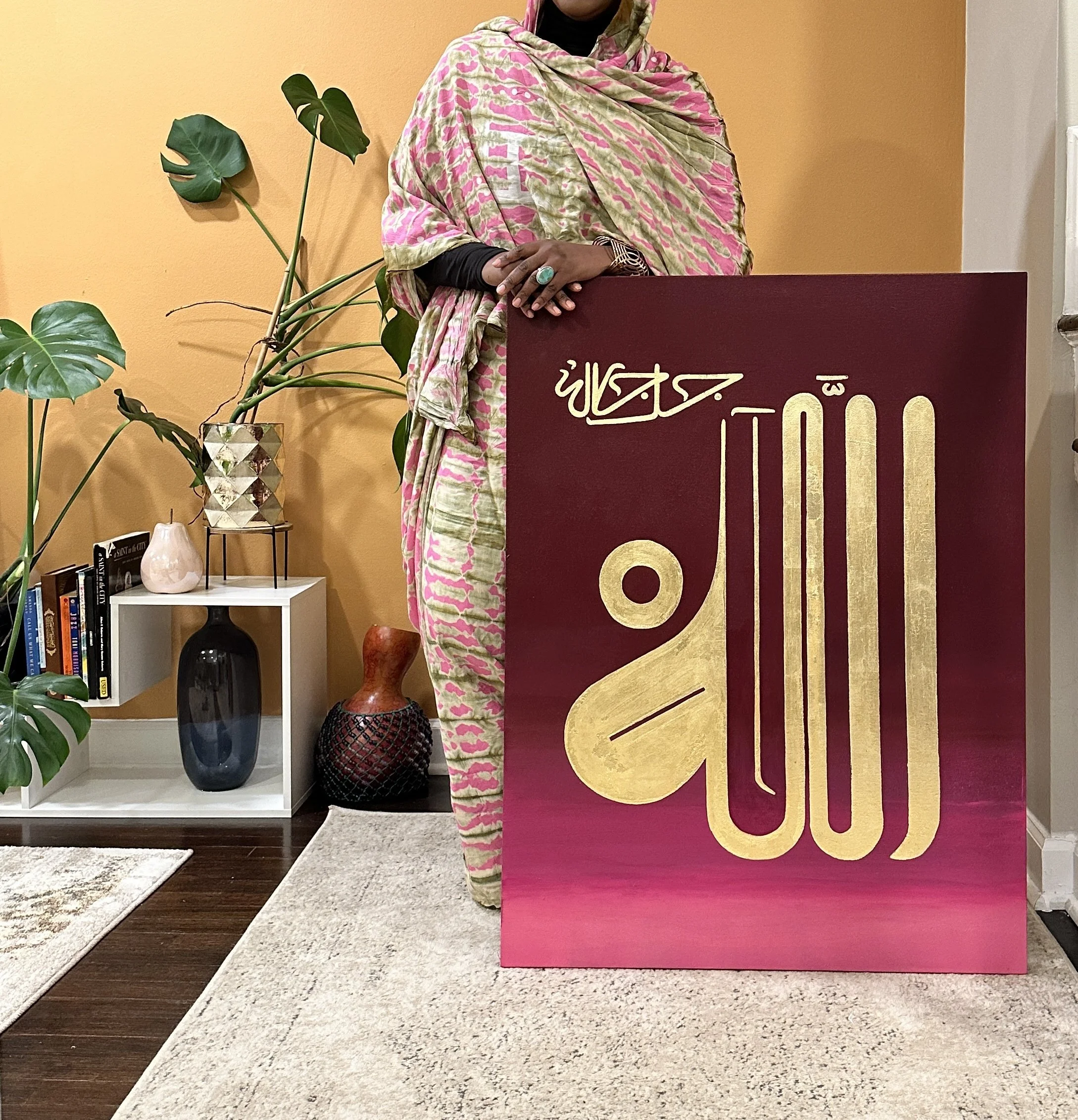 A woman dressed in traditional attire, standing next to a large maroon and gold Islamic calligraphy artwork, inside a modern room decorated with houseplants, books, and decorative vases.