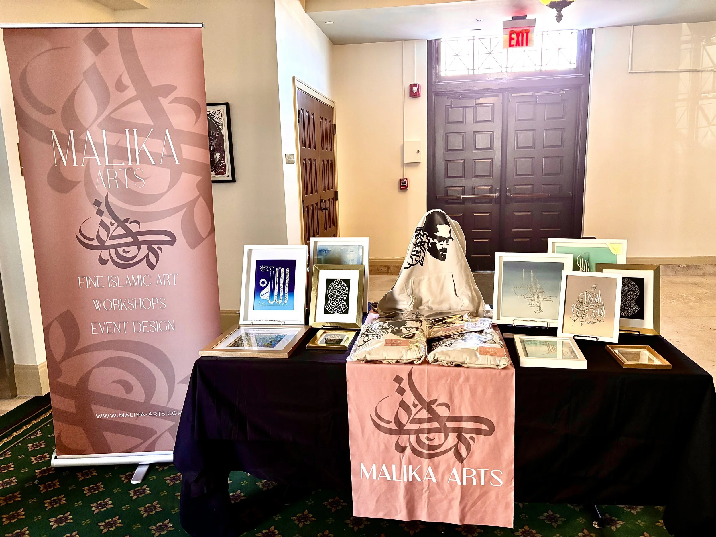 An art display booth for Malika Arts featuring framed Islamic art pieces, a mannequin with Islamic calligraphy, and a pink banner with the company logo and information.
