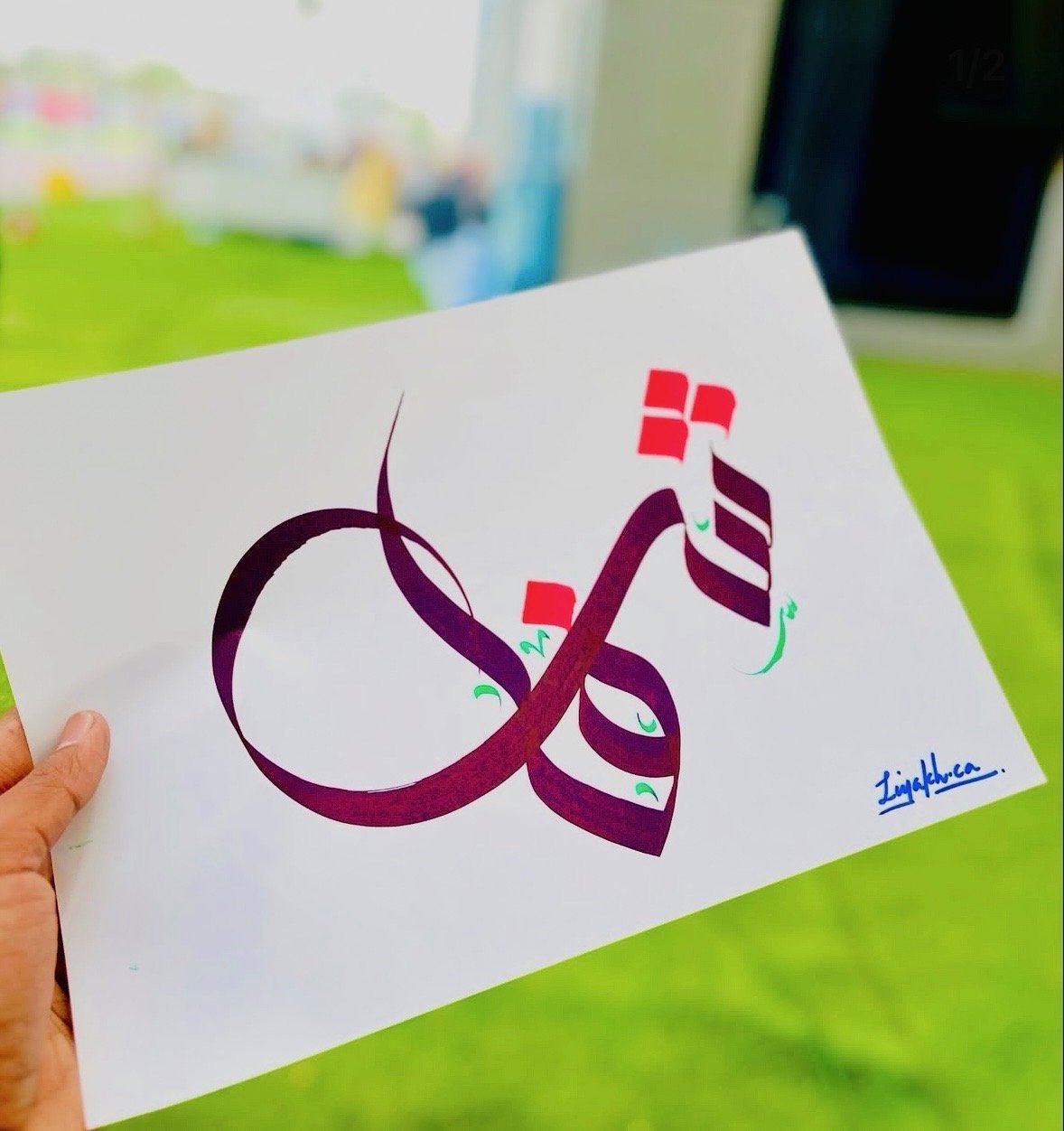 A hand holding a piece of paper with stylized Islamic calligraphy, with colorful accents and a signature in blue ink at the bottom right corner, all set against a blurred background of a green surface and shades of white and blue.