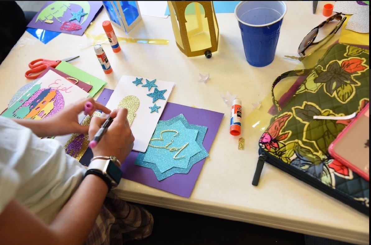 A person is crafting Islamic greeting cards with glitter and foam stickers. The table has glue sticks, scissors, paper, and a colorful fabric bag. There is also a blue cup, sunglasses, and a yellow paper lantern on the table.
